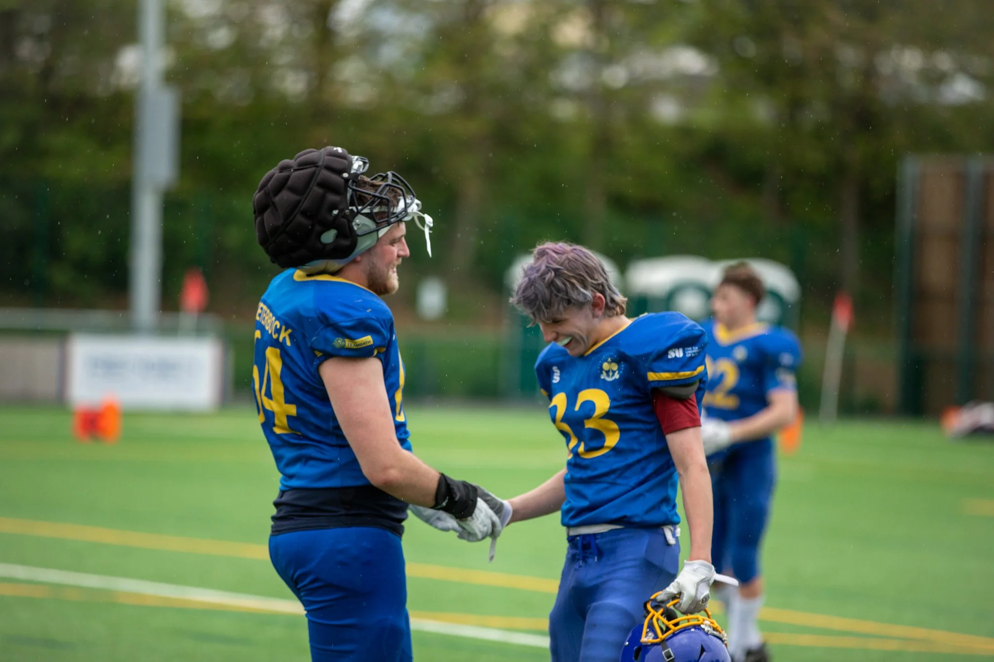 Two football players wearing blue jerseys shake hands on the field, with one wearing a helmet. Other players are visible in the background, and the scene takes place outdoors during the daytime.