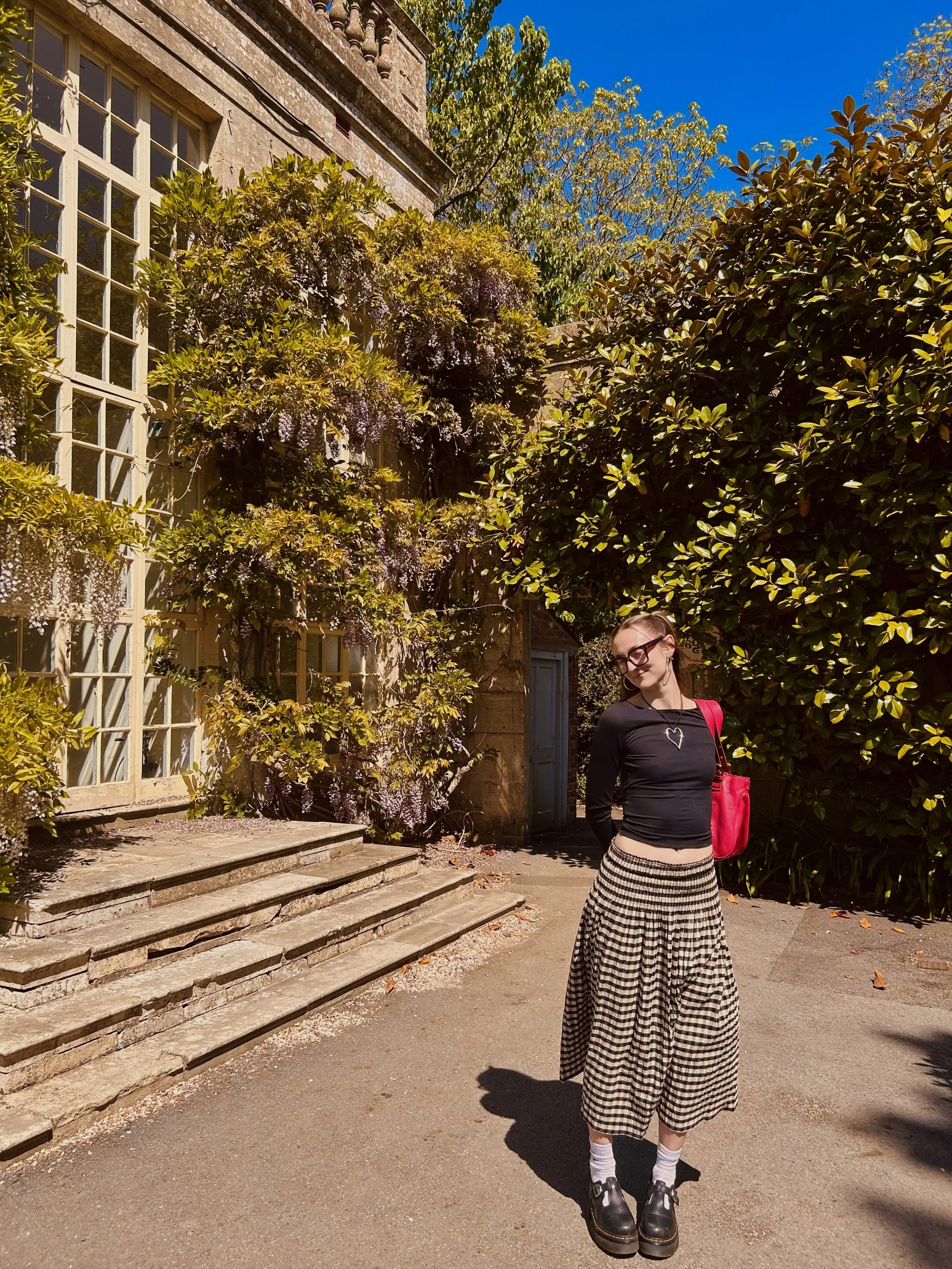 A woman standing outdoors on a sunny day, wearing a black long-sleeve shirt, checkered wide-leg pants, and platform shoes, next to a large green bush and a stone staircase leading up to a building with large windowpanes, trees, and a clear blue sky in the background.