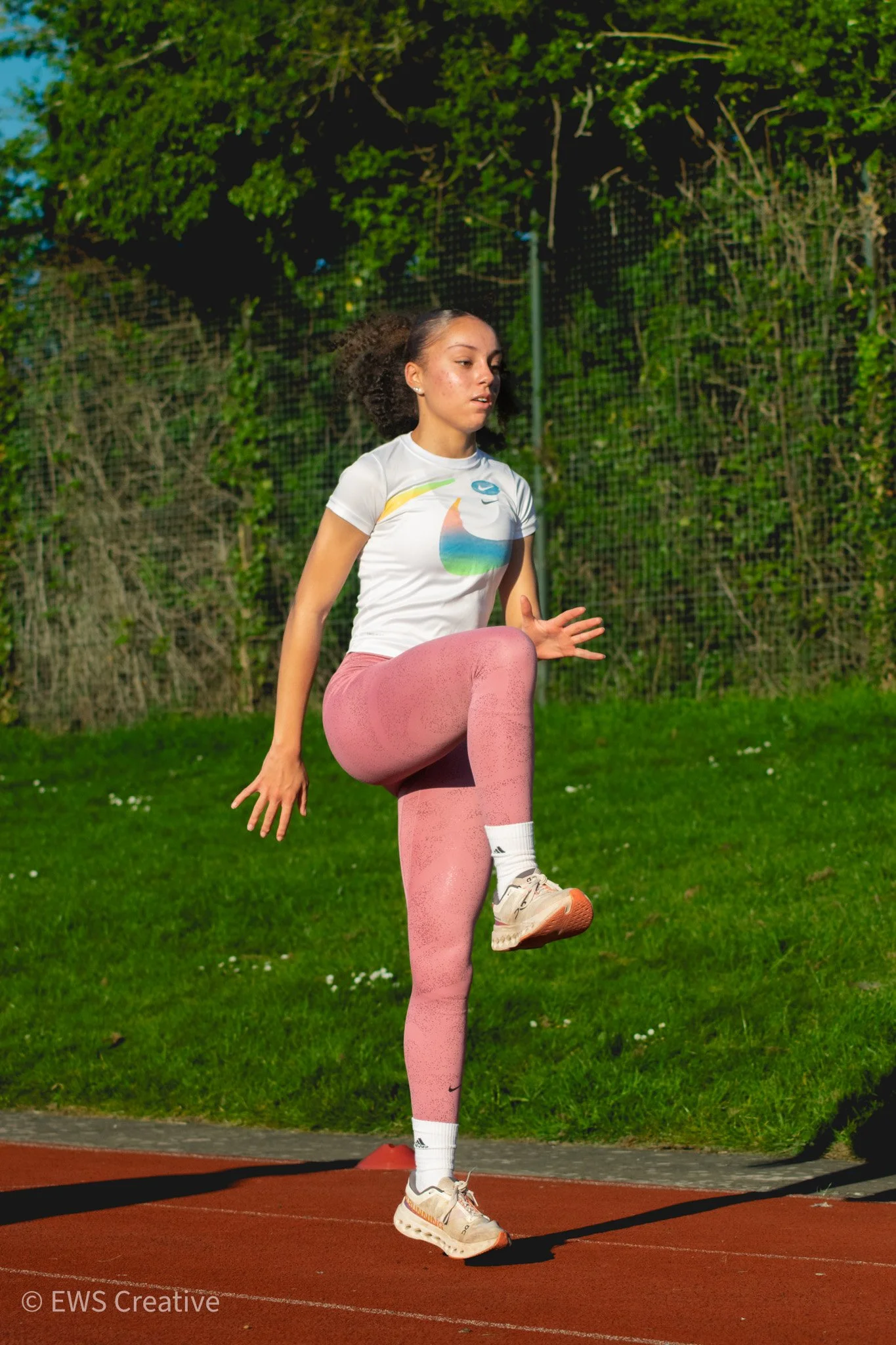 A young woman with curly hair tying her shoelace on an outdoor running track, wearing pink leggings, a white athletic shirt with a colorful print, and white sneakers. Green trees and a fence are in the background during daylight.