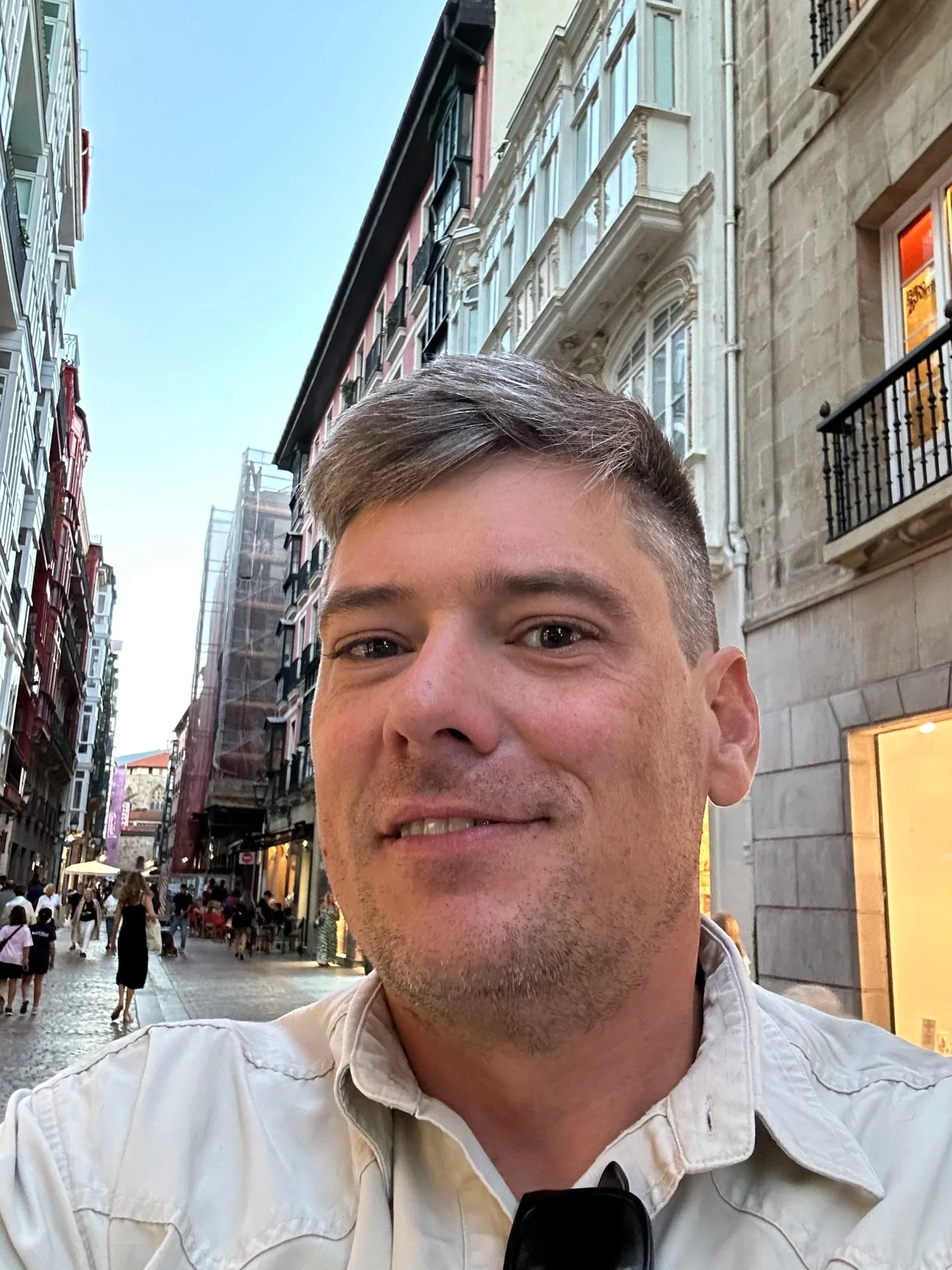 A young man taking a selfie on a busy European city street with historic buildings, outdoor cafes, and people walking in the background during the evening.