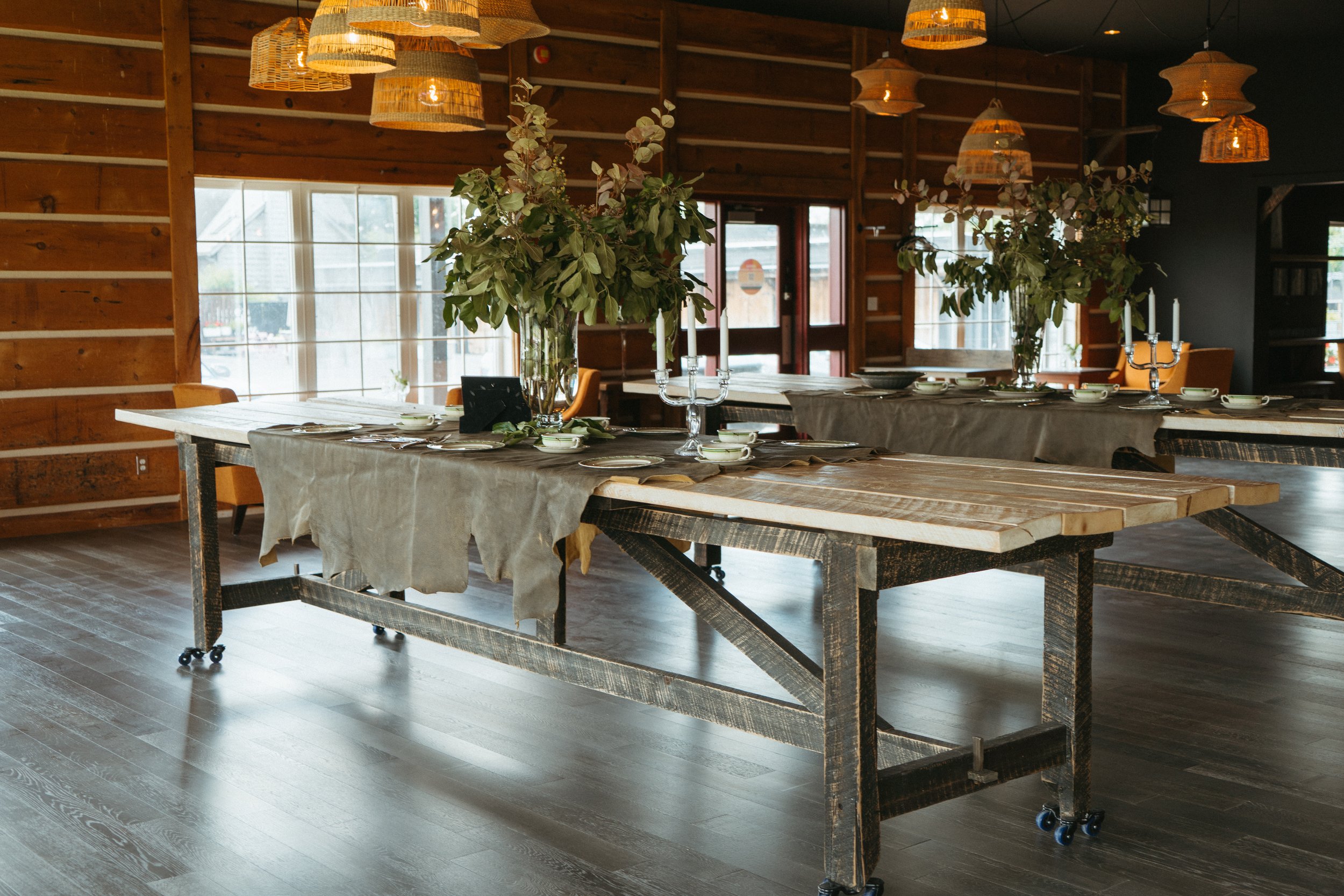 A rustic dining room with a long wooden table covered with a beige tablecloth, set with white plates and teal cups, adorned with large vases of green foliage, and surrounded by wooden chairs. Pendant lights hang from a dark ceiling, and large windows let in natural light.