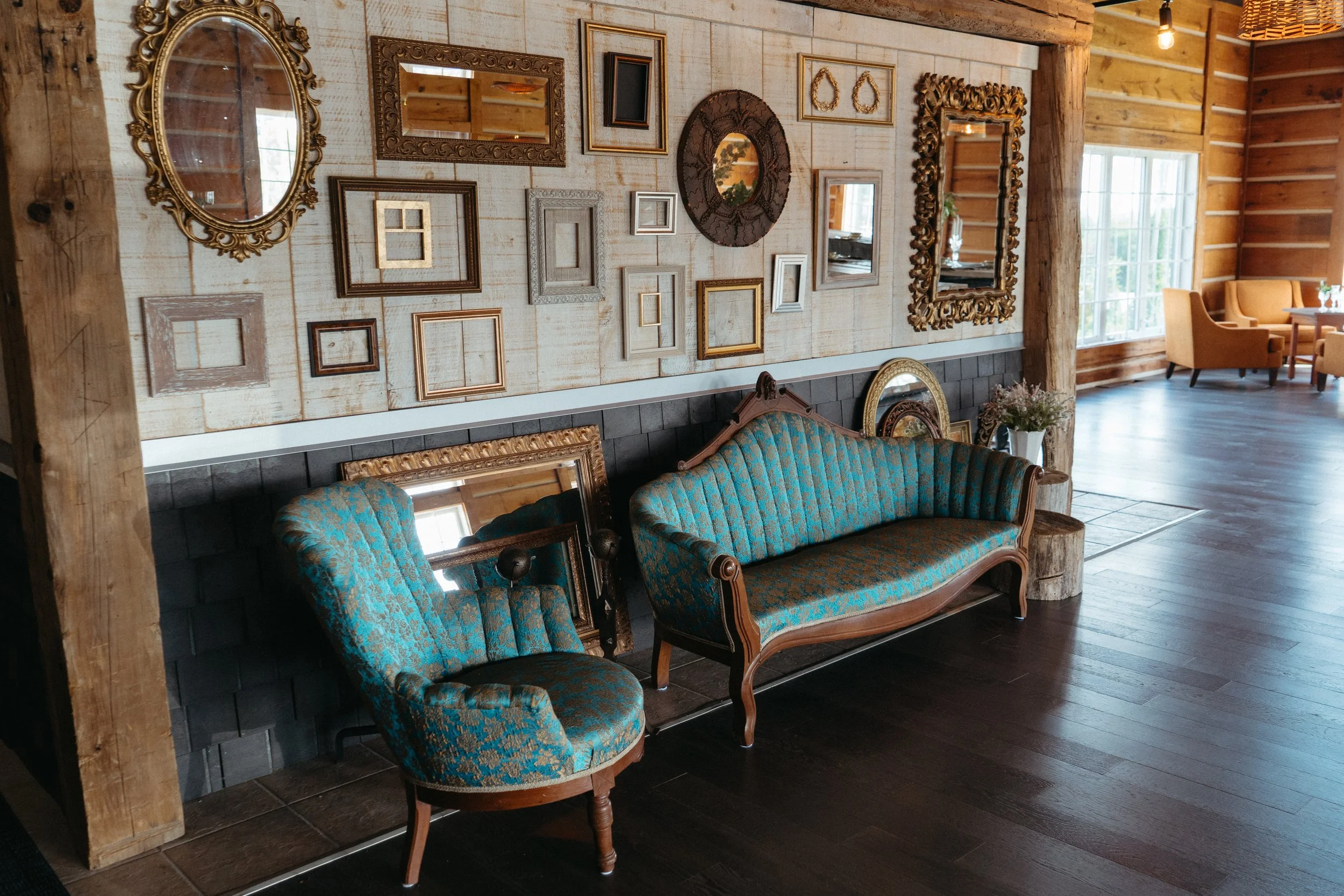Interior of a rustic room with a decorated wall of empty picture frames and mirrors, vintage blue upholstered sofa, wooden beams, a window with sunlight, and a cozy seating area.