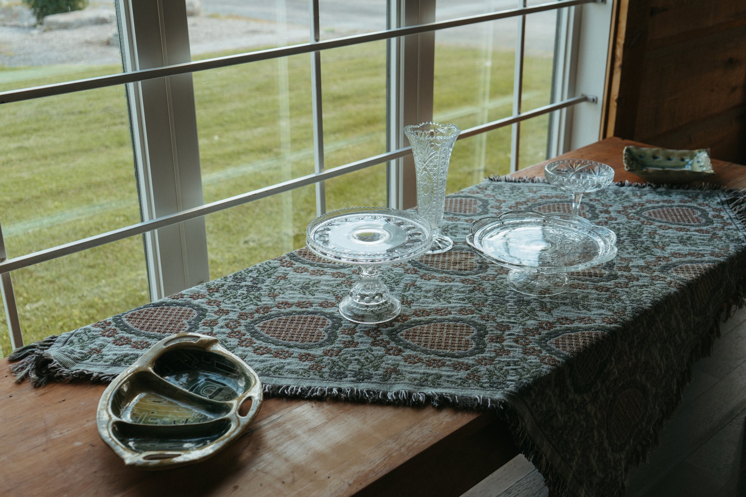 A wooden table with a patterned cloth, set with various glassware including a tall vase, a bowl, and a cake stand, near a window overlooking a grassy yard.