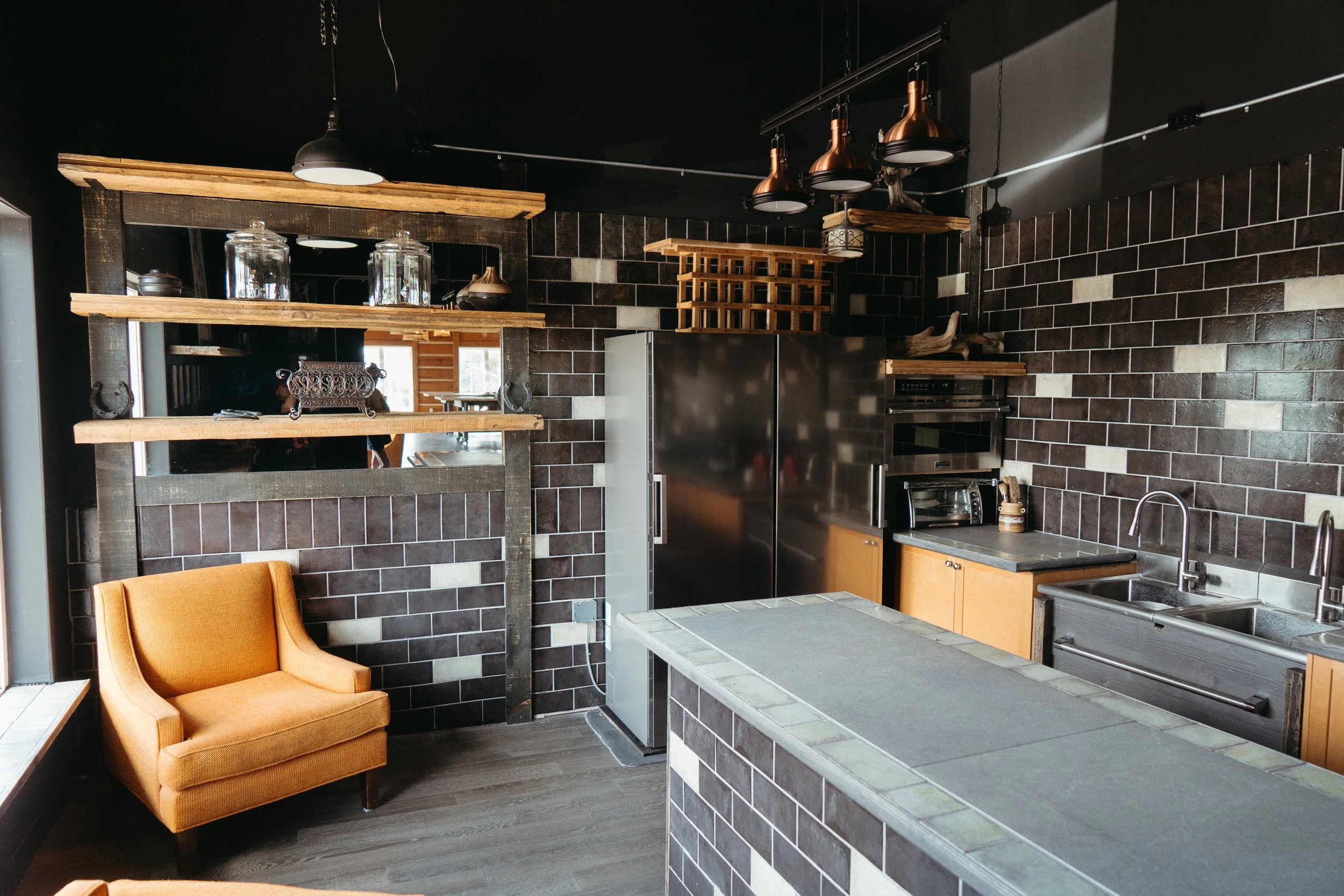 A modern kitchen with dark brick walls, wooden shelves, a stainless steel refrigerator, microwave, and sink. There are copper pendant lights hanging from the ceiling and an orange armchair in the foreground.