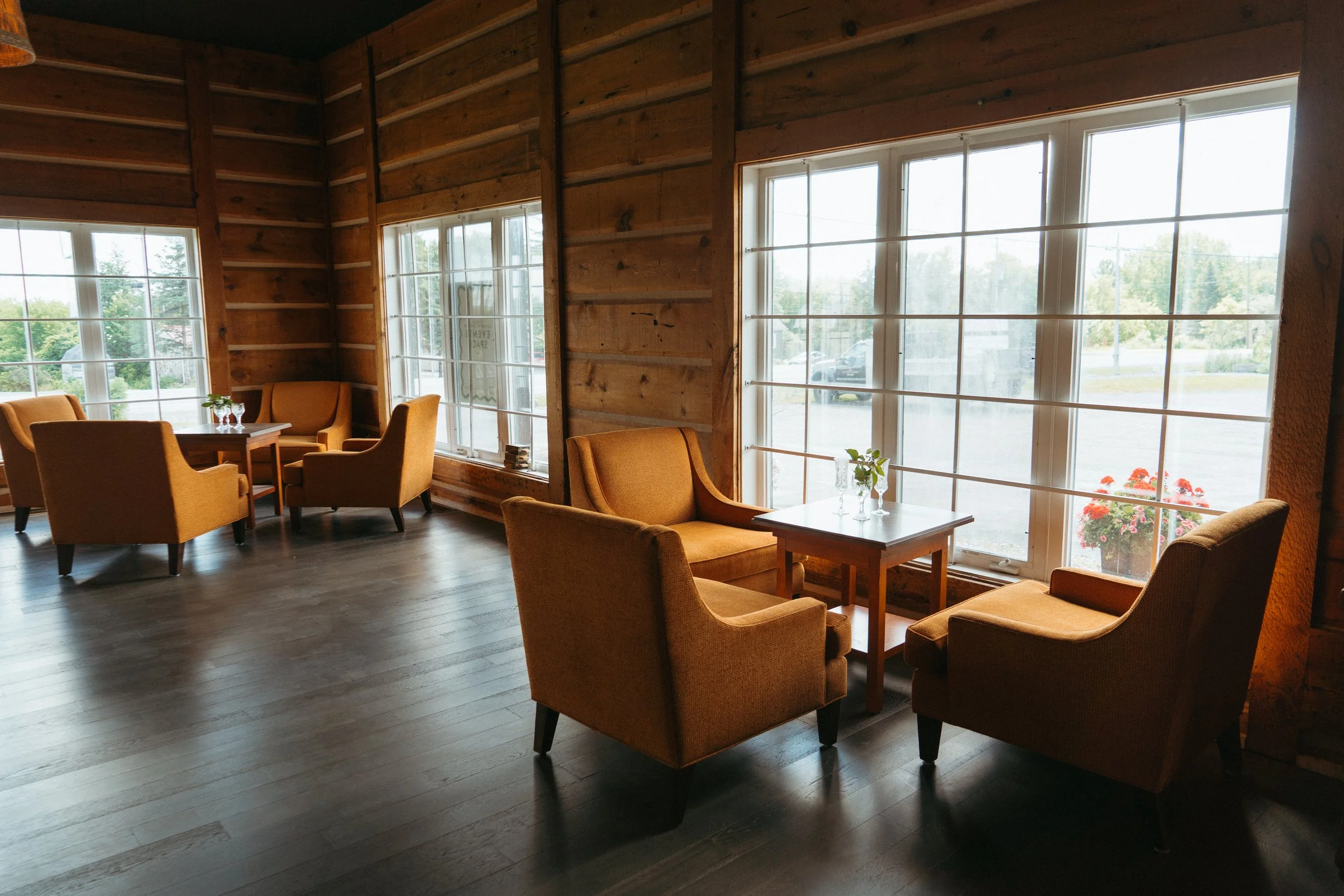 Interior of a cozy room with large windows, wooden wall paneling, and orange armchairs arranged around small tables with flowers, providing a view of greenery outside.