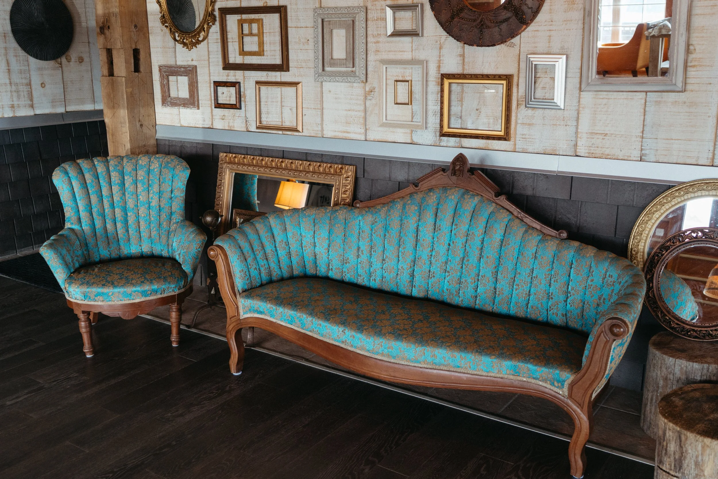 Vintage sitting area with a patterned blue sofa and matching armchair, framed mirrors and empty picture frames on a rustic wall.