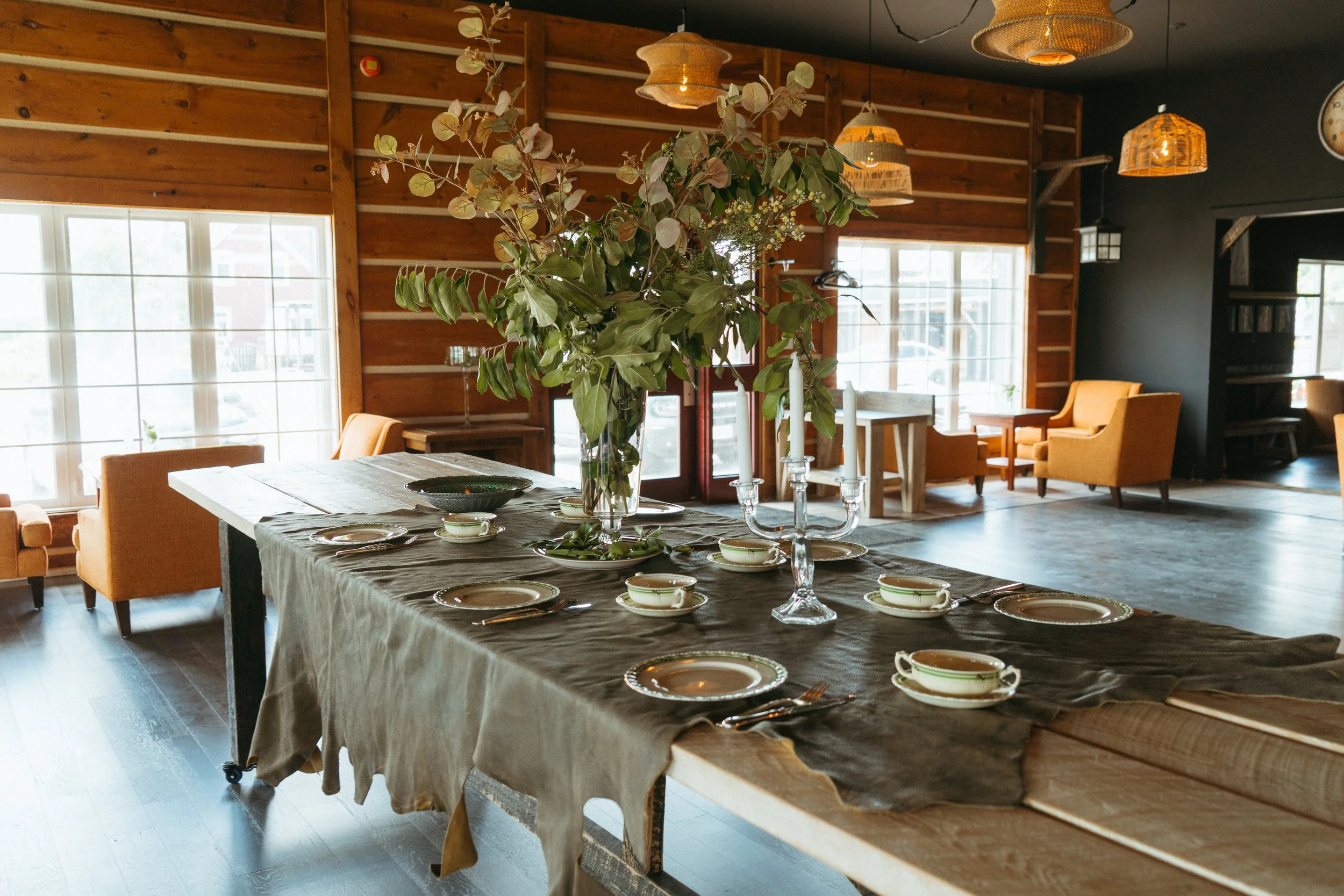 A dining area with a wooden table set with plates and cutlery, a large flower arrangement centerpiece, and a background with yellow armchairs, wooden wall panels, and windows letting in natural light.
