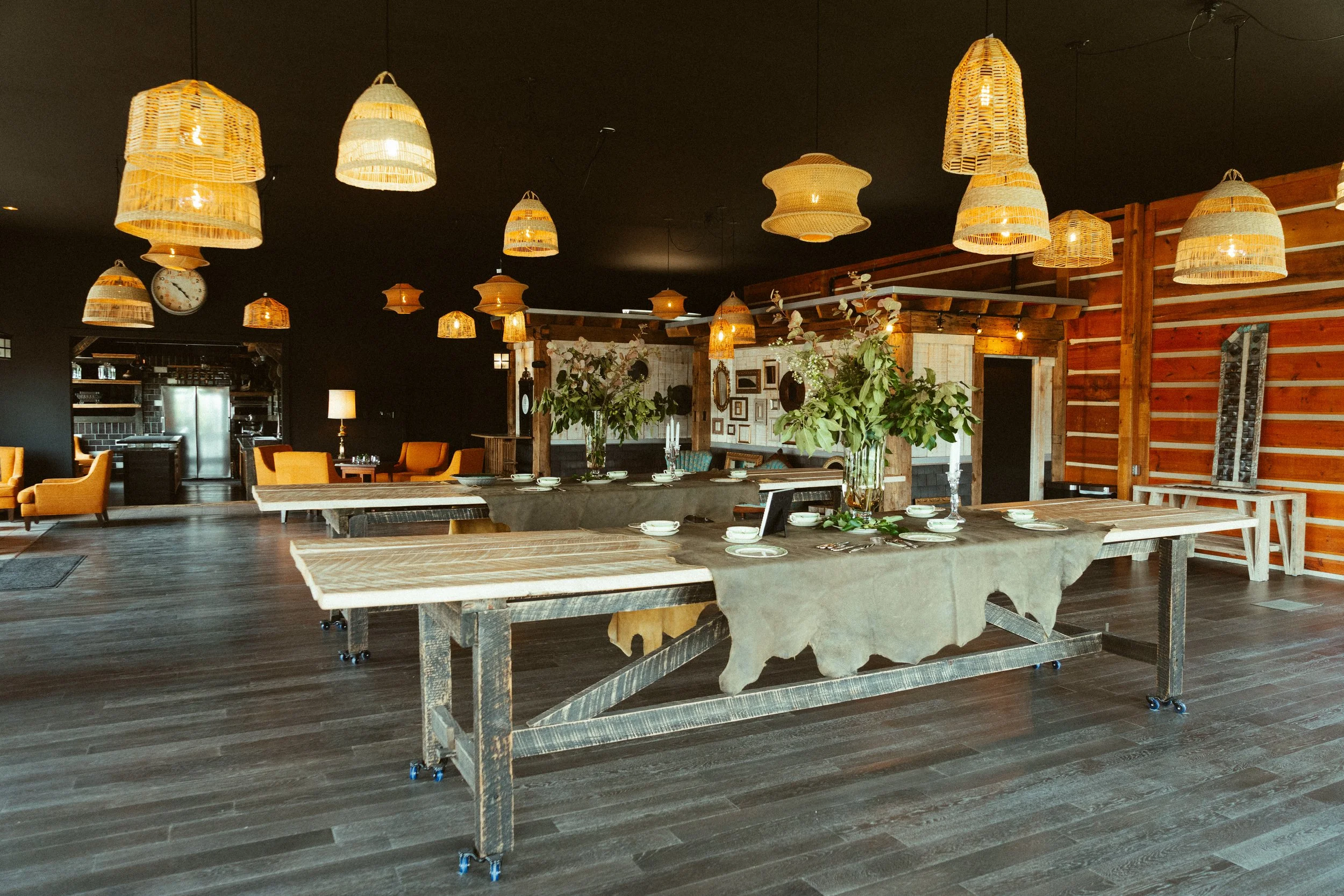A rustic dining space with hanging wicker lamps, a large table set with plates and silverware, and a central floral arrangement, with wood panel walls and a dark ceiling.