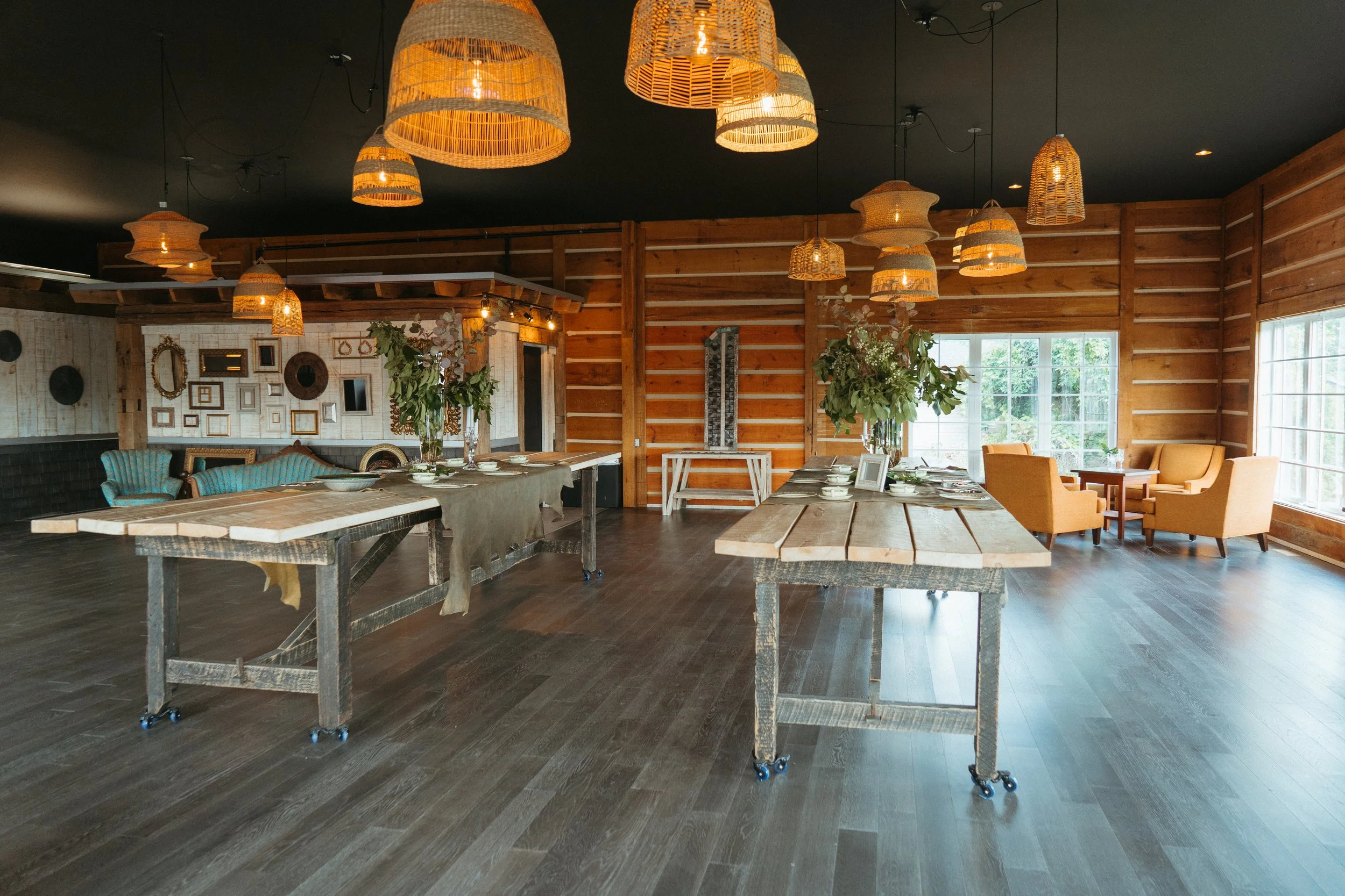A rustic indoor space with wooden walls, large windows, and multiple woven pendant lights hanging from a black ceiling. There are tables set with white dishes and floral centerpieces, and cozy seating areas with chairs and sofas.