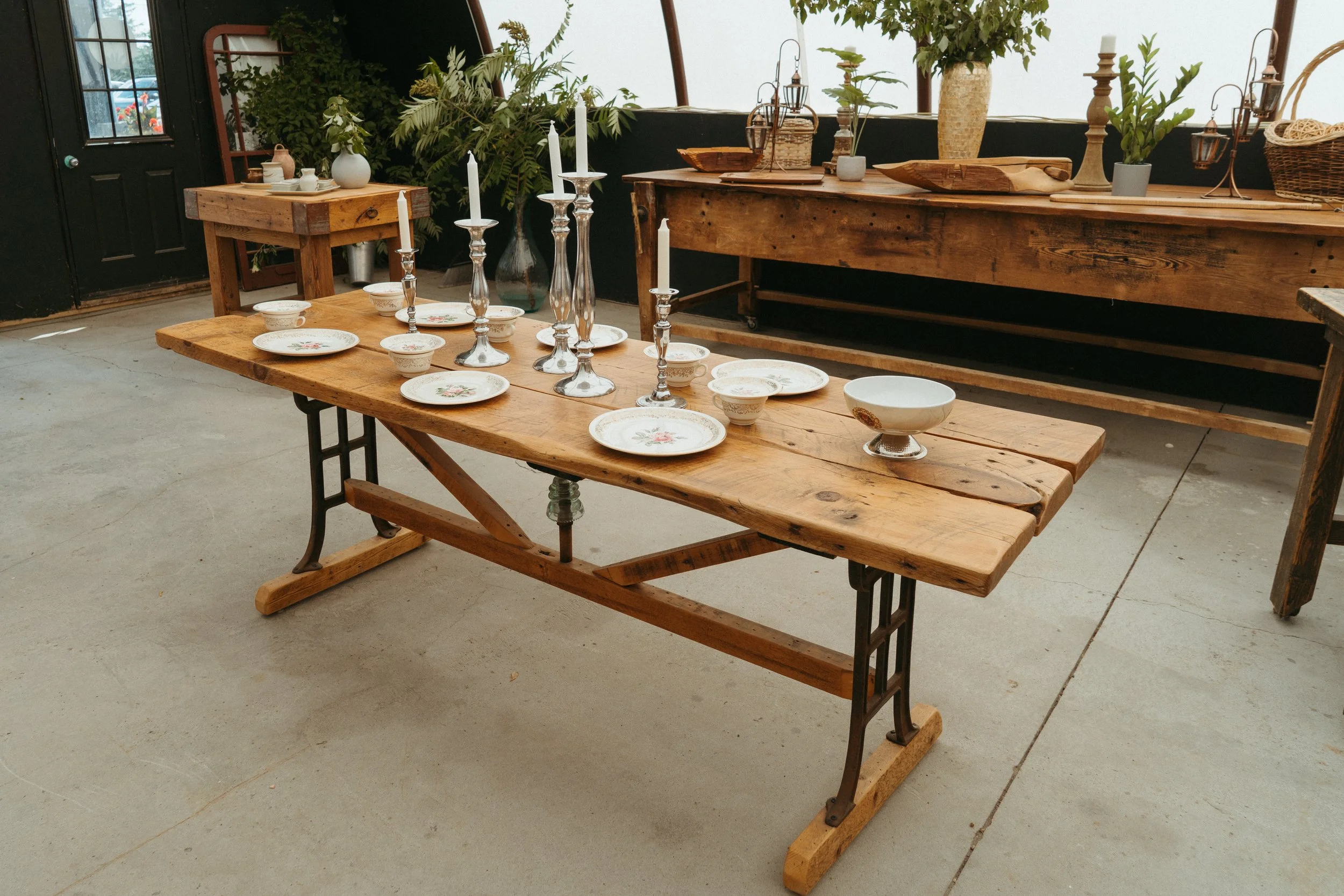 A wooden dining table set on a concrete floor, decorated with plates, bowls, and tall candlesticks, in a rustic, greenhouse-like setting with potted plants and baskets on the side.