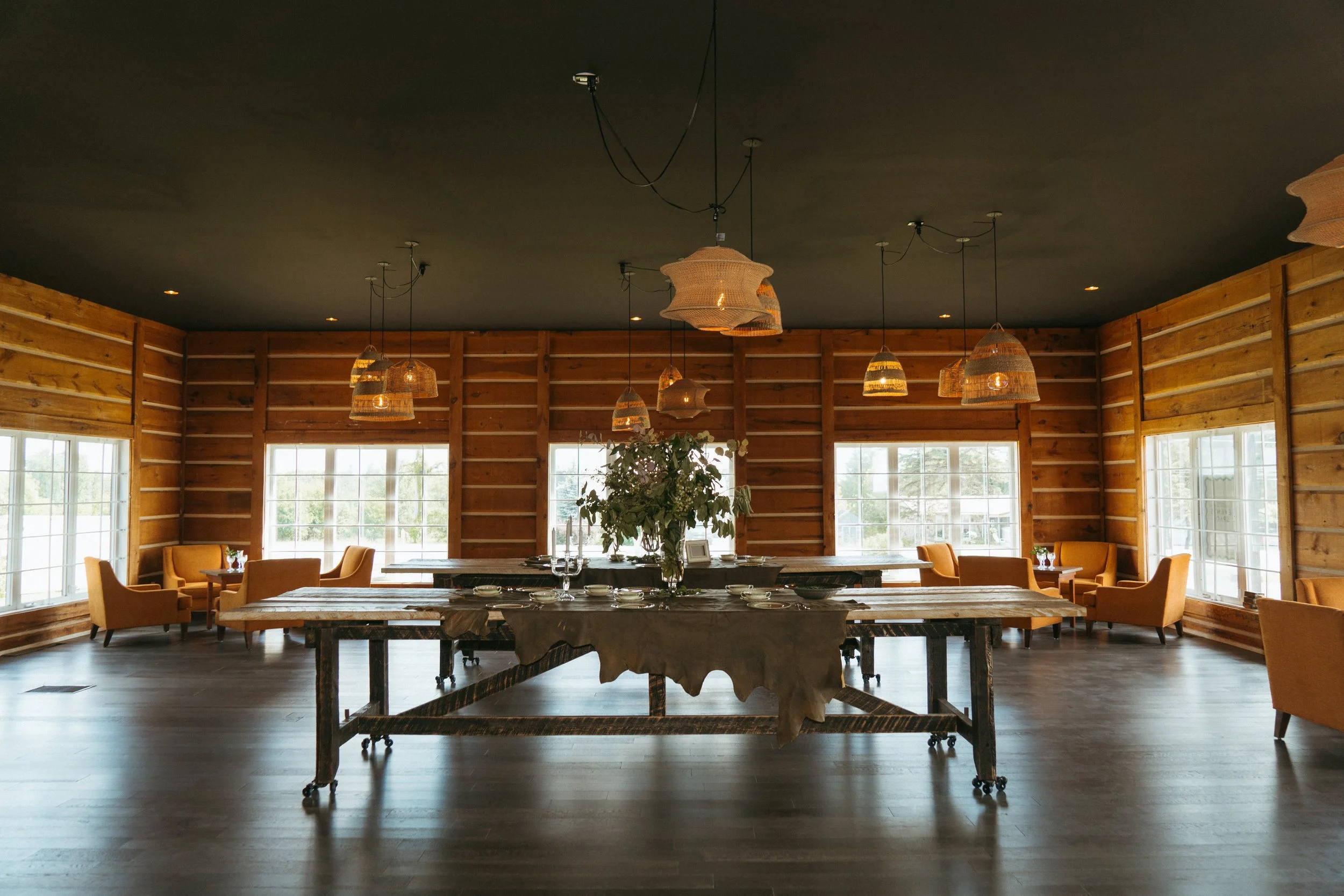 Interior of a rustic dining area with wooden walls, large windows, hanging woven pendant lights, yellow armchairs around small tables, and a large central wooden table with a floral centerpiece.