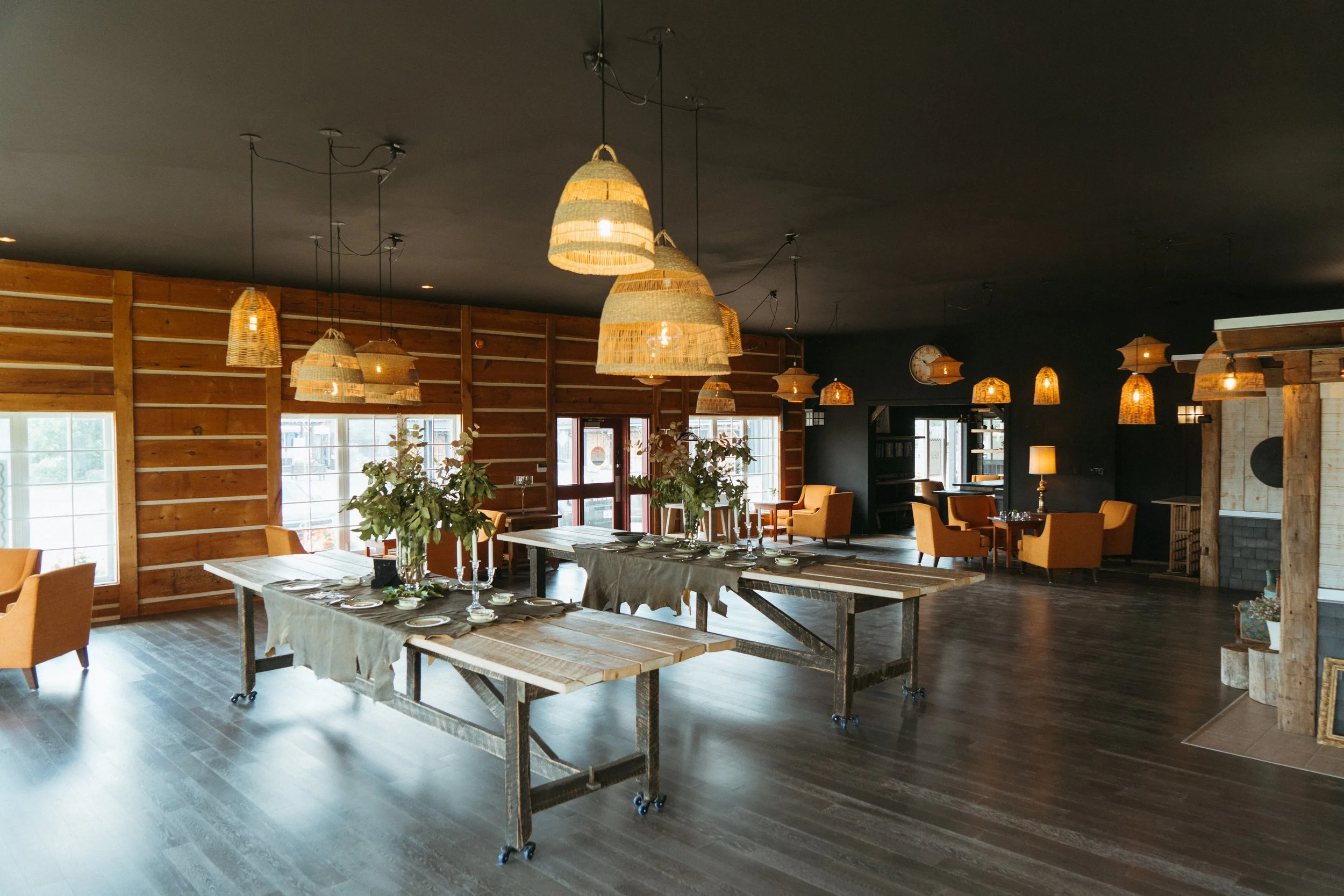 Interior of a rustic-style event space or restaurant with wooden walls, multiple woven pendant lights hanging from the dark ceiling, and a long table set with dishes, glasses, and candles. There are additional seating areas with orange chairs and tab