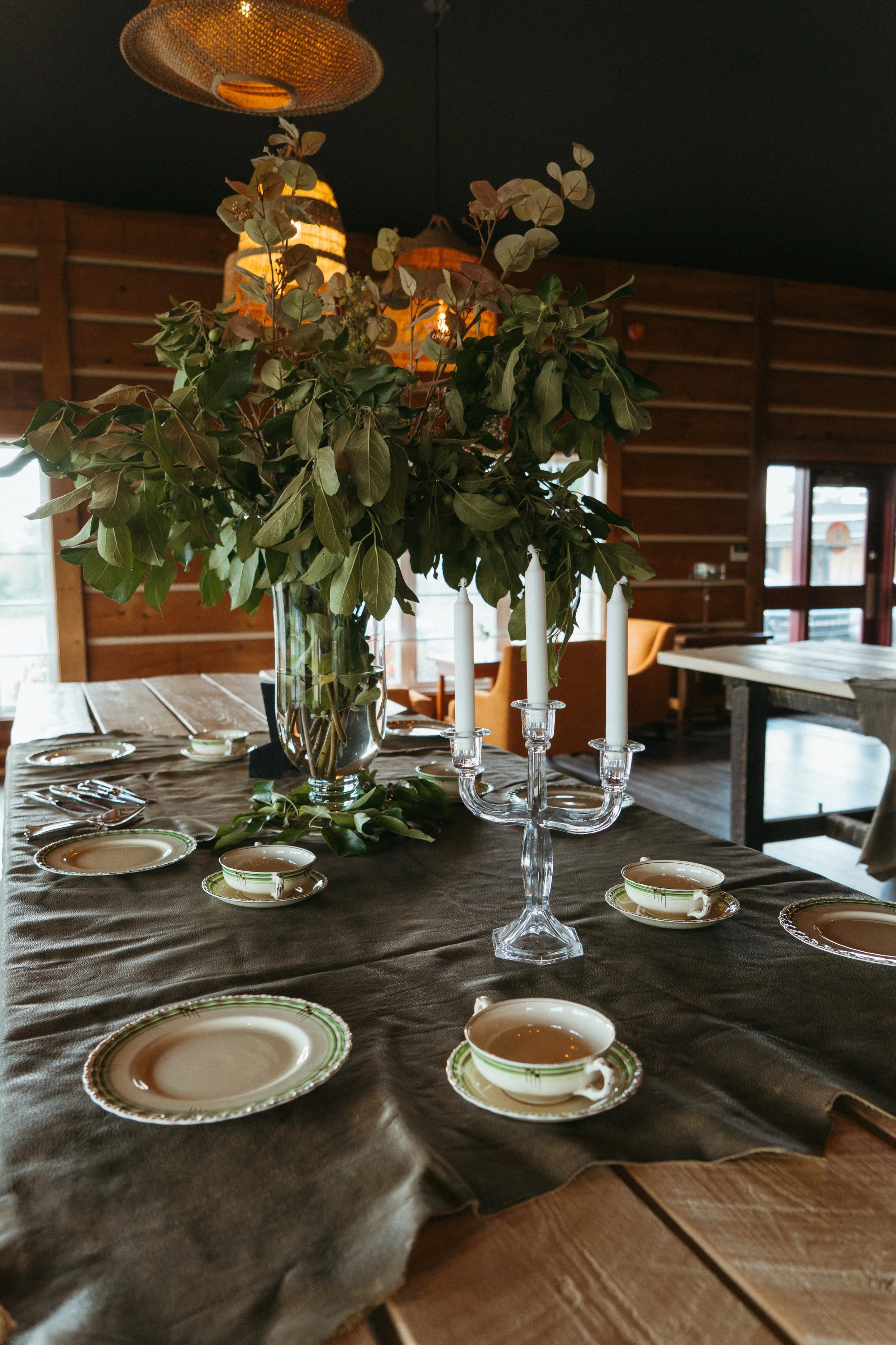 A dining table set with vintage china, a gray tablecloth, a glass candelabrum, and a large floral arrangement with eucalyptus leaves. The background shows a wood-paneled room with large windows and hanging lamps.