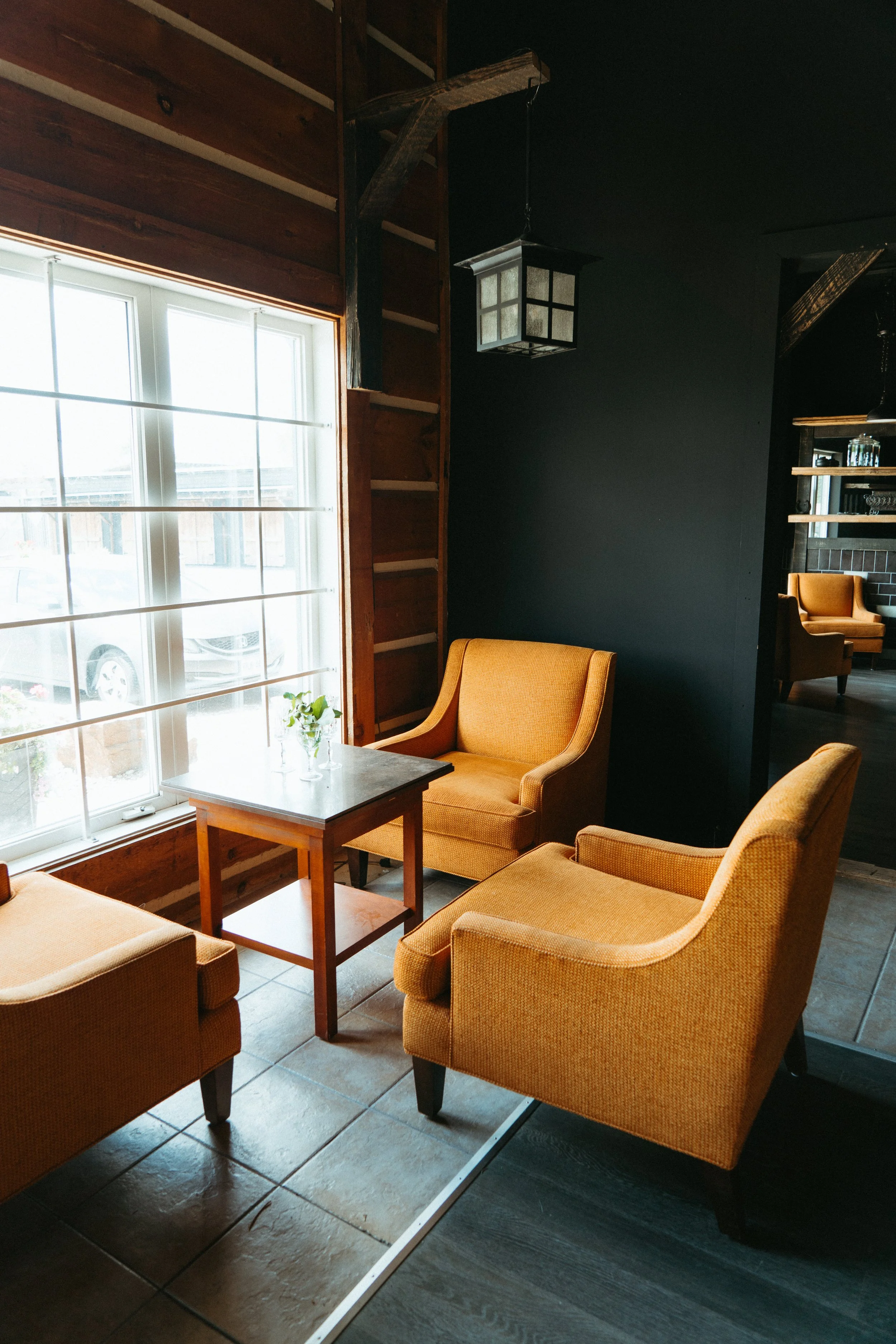 Interior of a cozy cafe with mustard yellow armchairs around a small wooden table, large window with white frames letting in natural light, dark walls, and a wooden ceiling with exposed beams and pendant lamps.