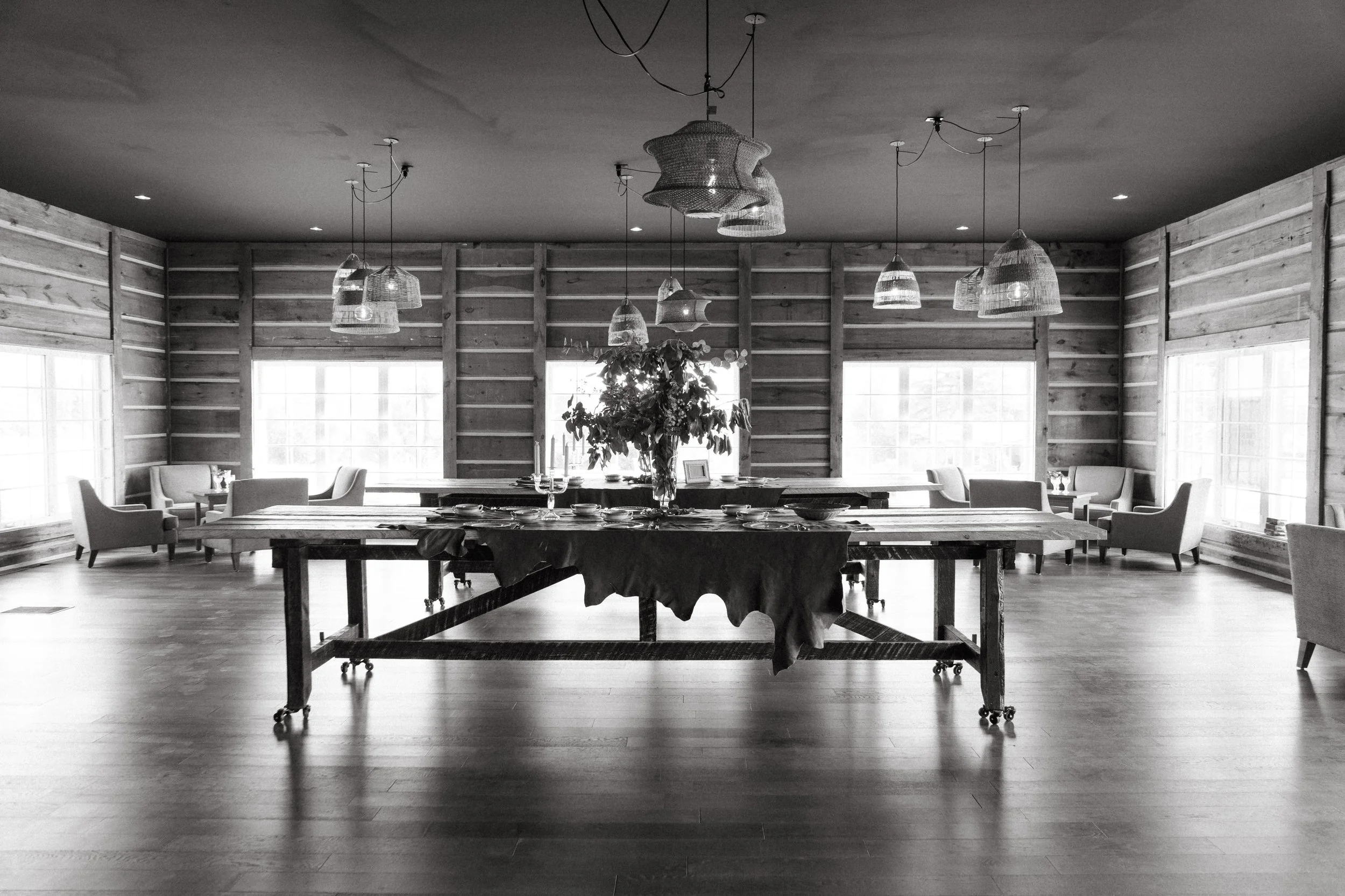 A spacious dining room with a long wooden table covered with a tablecloth, surrounded by chairs. The room has wooden panel walls and large windows letting in natural light. Multiple hanging pendant lights decorate the ceiling, and there is a large fl