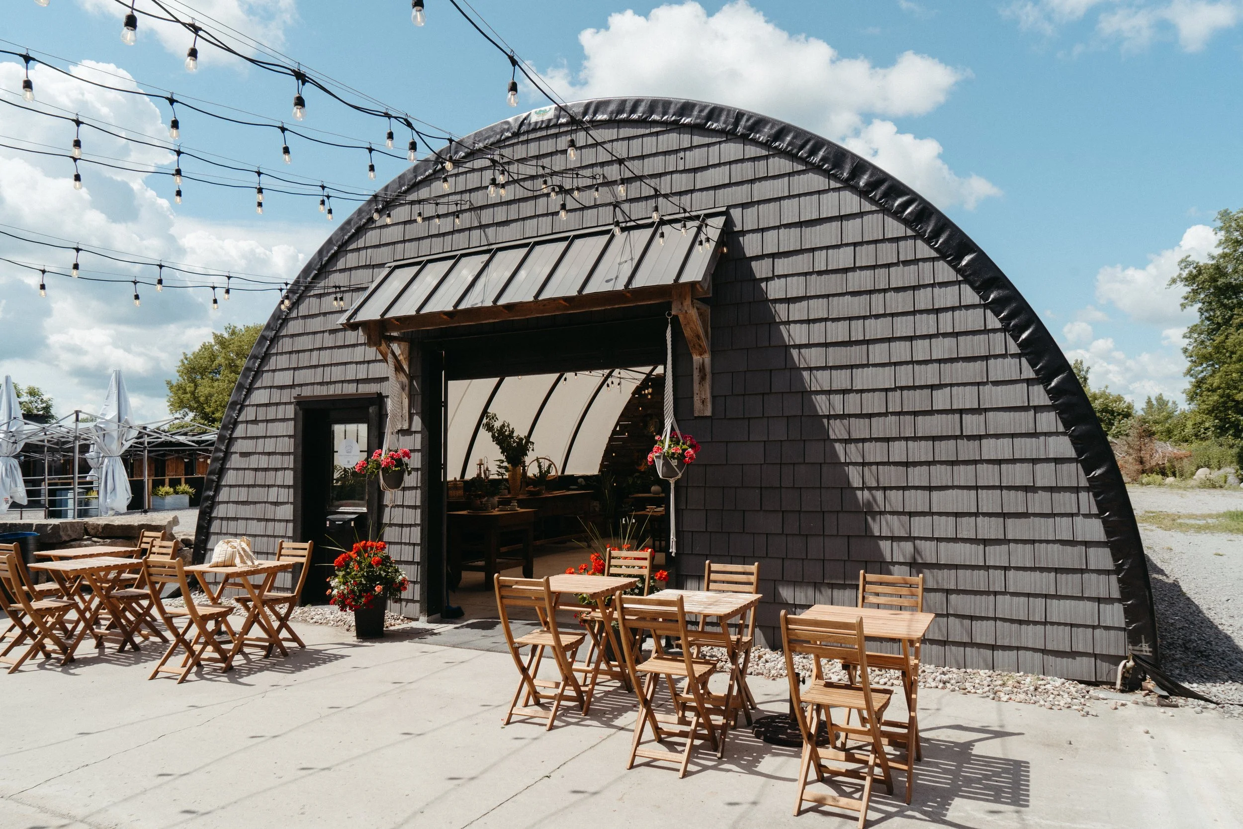 Outdoor patio with wooden tables and chairs set up in front of a rustic barn-style building with dark gray shingle siding. String lights are hung above the seating area. There are potted flowers near the entrance, and trees and umbrellas are visible 