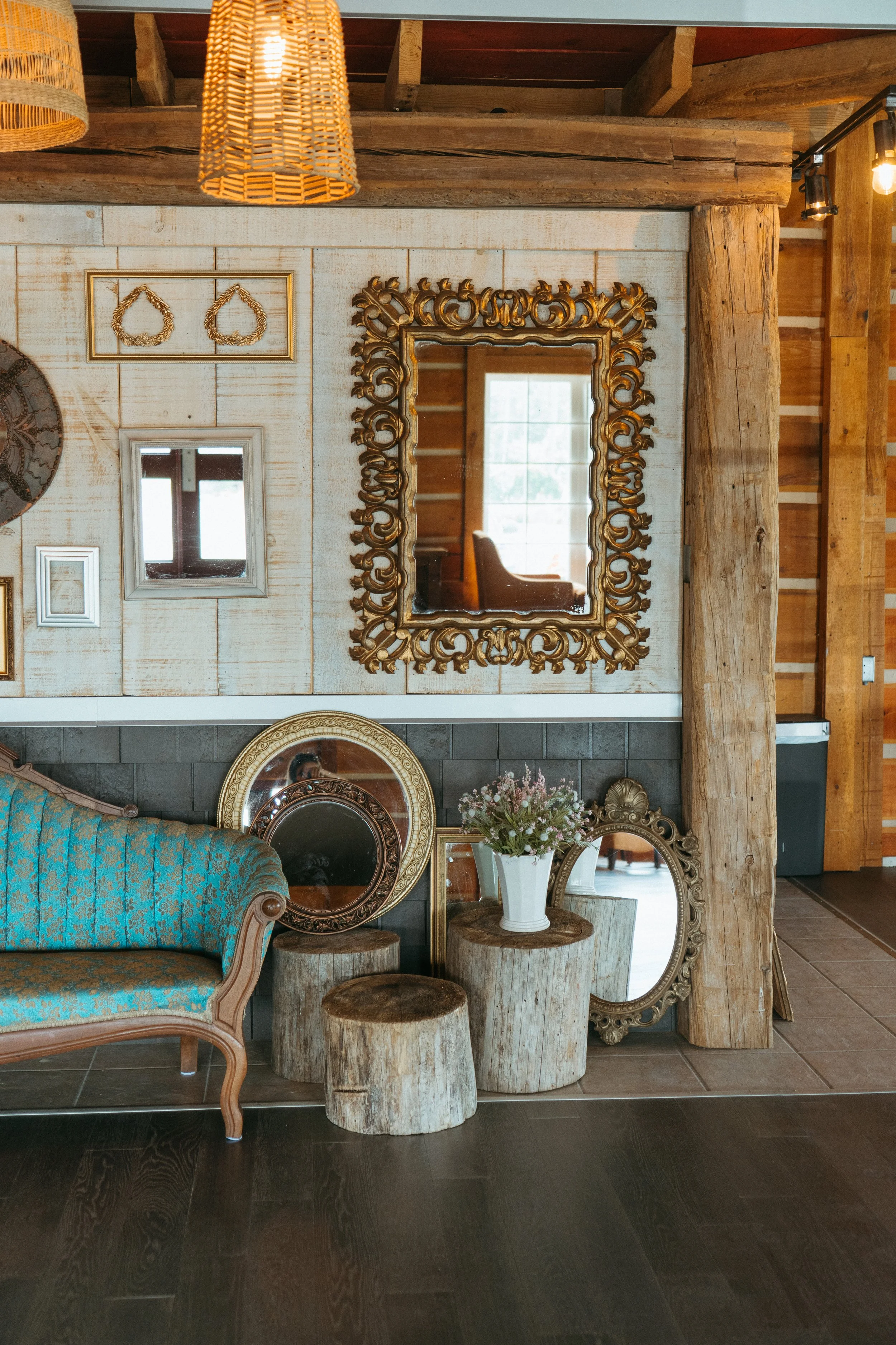 Living room with rustic decor, including a vintage teal armchair with wood trim, a white vase with flowers, three wooden logs, and a large ornate gold-framed mirror on the wall. The wall has decorative picture frames and a round decorative plate. The ceiling features wooden beams, with woven pendant lights hanging down. Sunlight streams through a window reflected in the mirror.