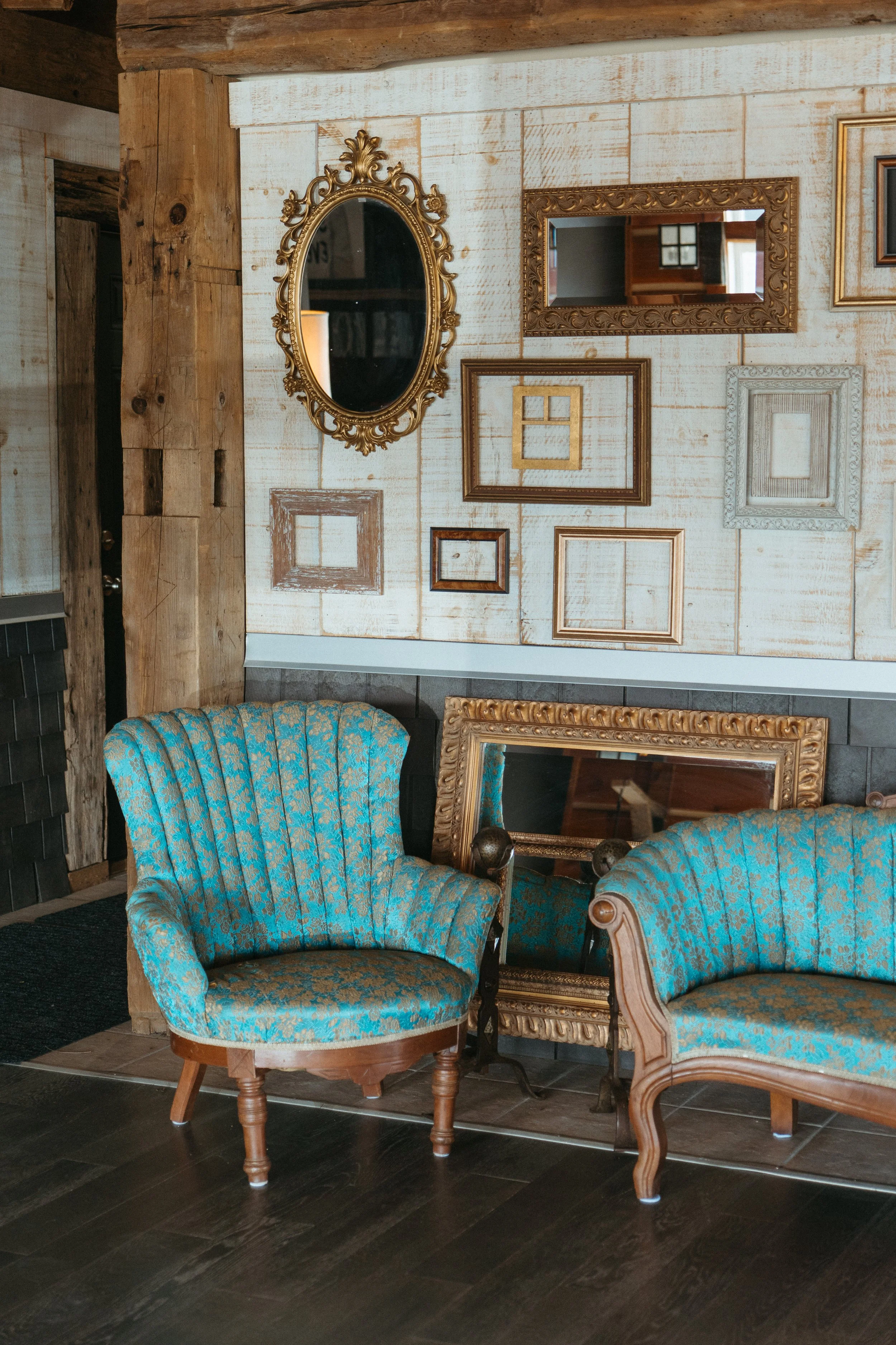 A cozy interior with vintage furniture, including two matching blue and gold ornate armchairs, set against a rustic wooden wall decorated with multiple empty picture frames and a gold-framed mirror.