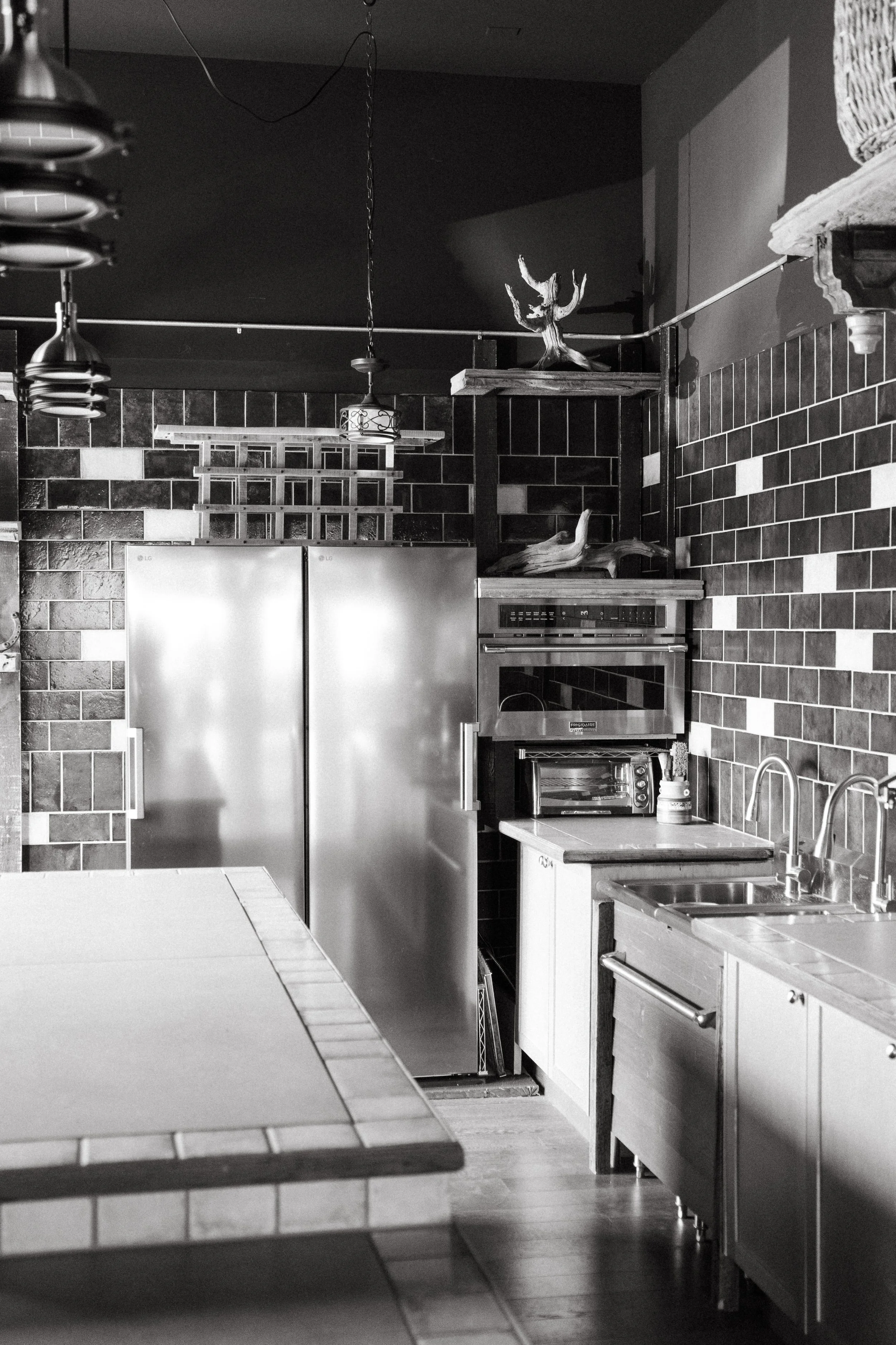 A black and white kitchen with brick-patterned tile walls, a stainless steel refrigerator, a built-in oven, a microwave, a sink, and wooden cabinets. There are decorative wooden branches on a shelf, and a ceiling light fixture is hanging from the cei