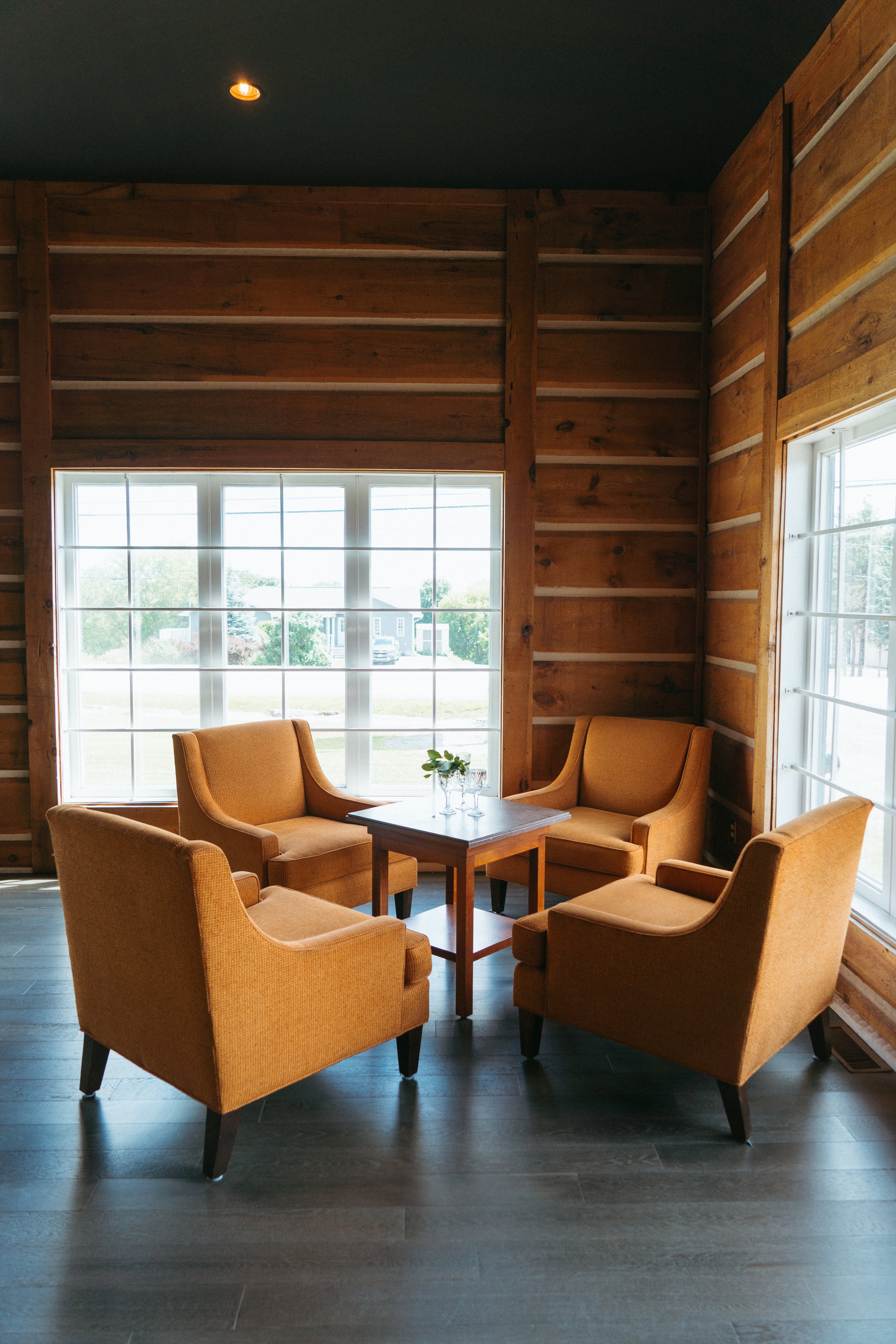 A cozy indoor seating area with six mustard-yellow armchairs arranged around a small wooden coffee table, set against wooden paneled walls and large windows letting in natural light.