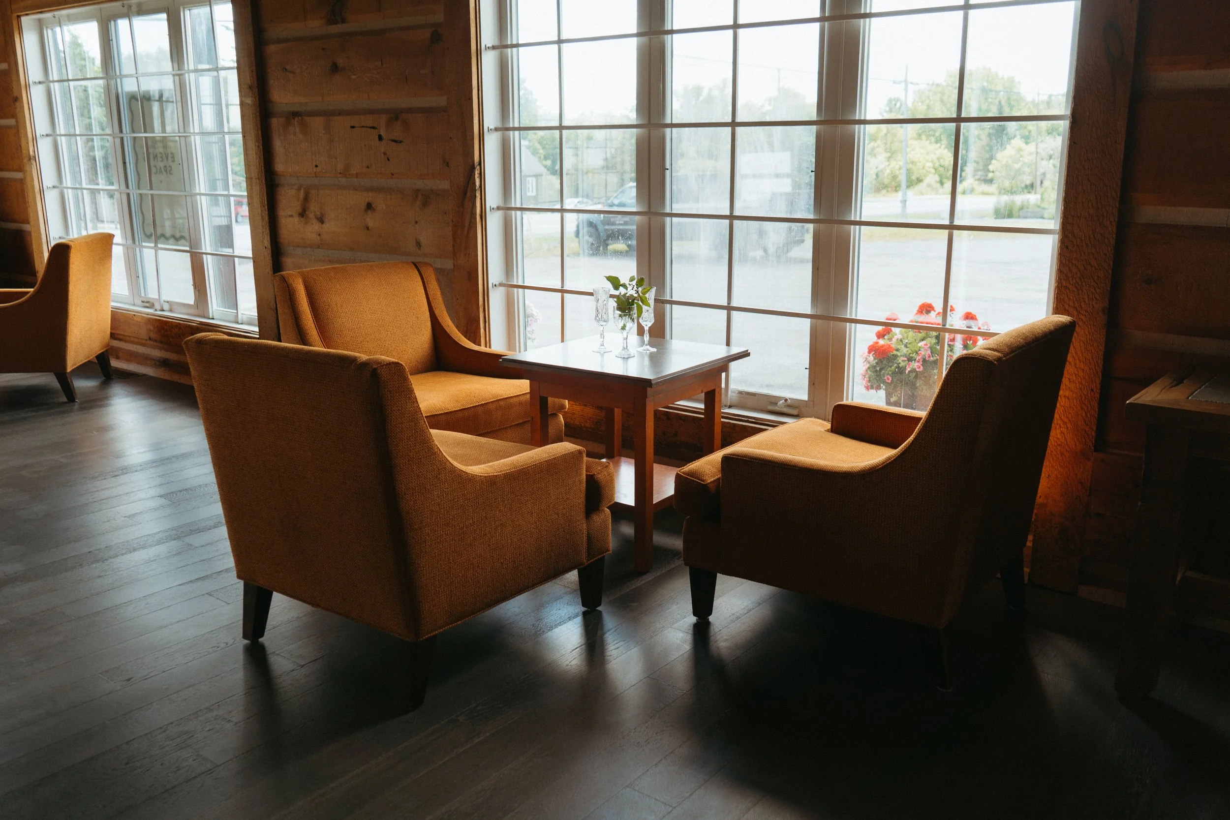 A cozy indoor seating area with comfortable armchairs around a small wooden table decorated with a small vase and glasses, situated near large windows with a view of the outdoor parking lot and greenery.