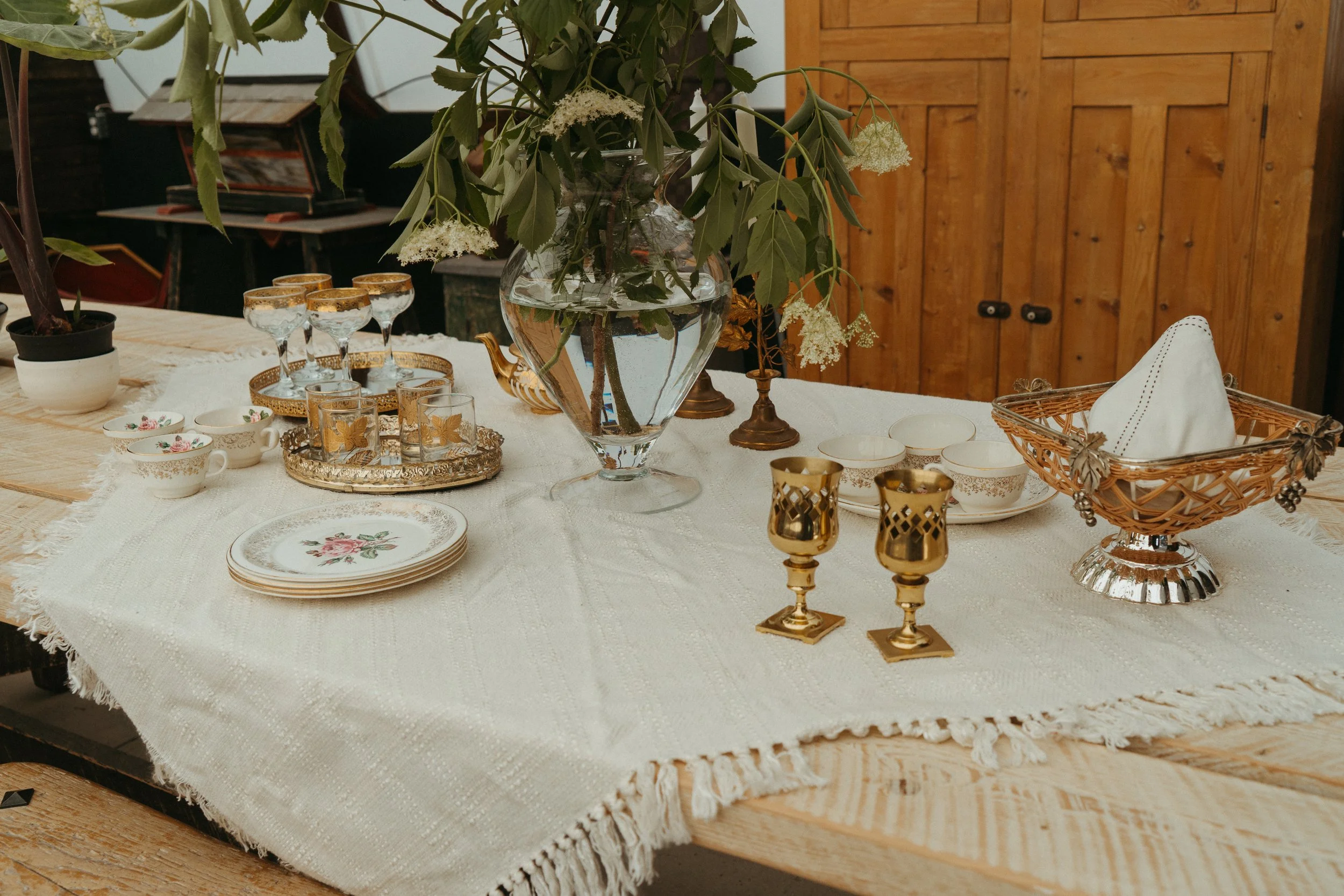 A dining table set with vintage china, gold-trimmed glassware, and various decorative items including a large glass vase with greenery and white flowers, brass candle holders, and a woven basket with a napkin, on a white tablecloth. In the background, there are wooden cabinets and a few chairs.