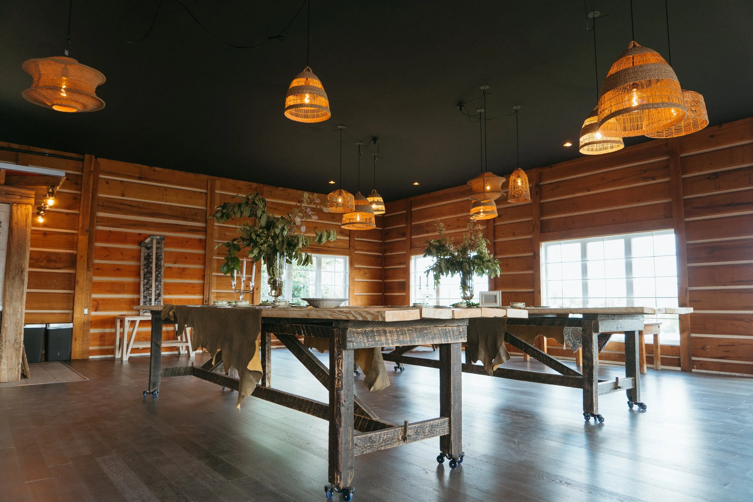 Rustic dining room with a long wooden table, floral centerpieces, and woven pendant lights hanging from a black ceiling, with wooden walls and large windows.