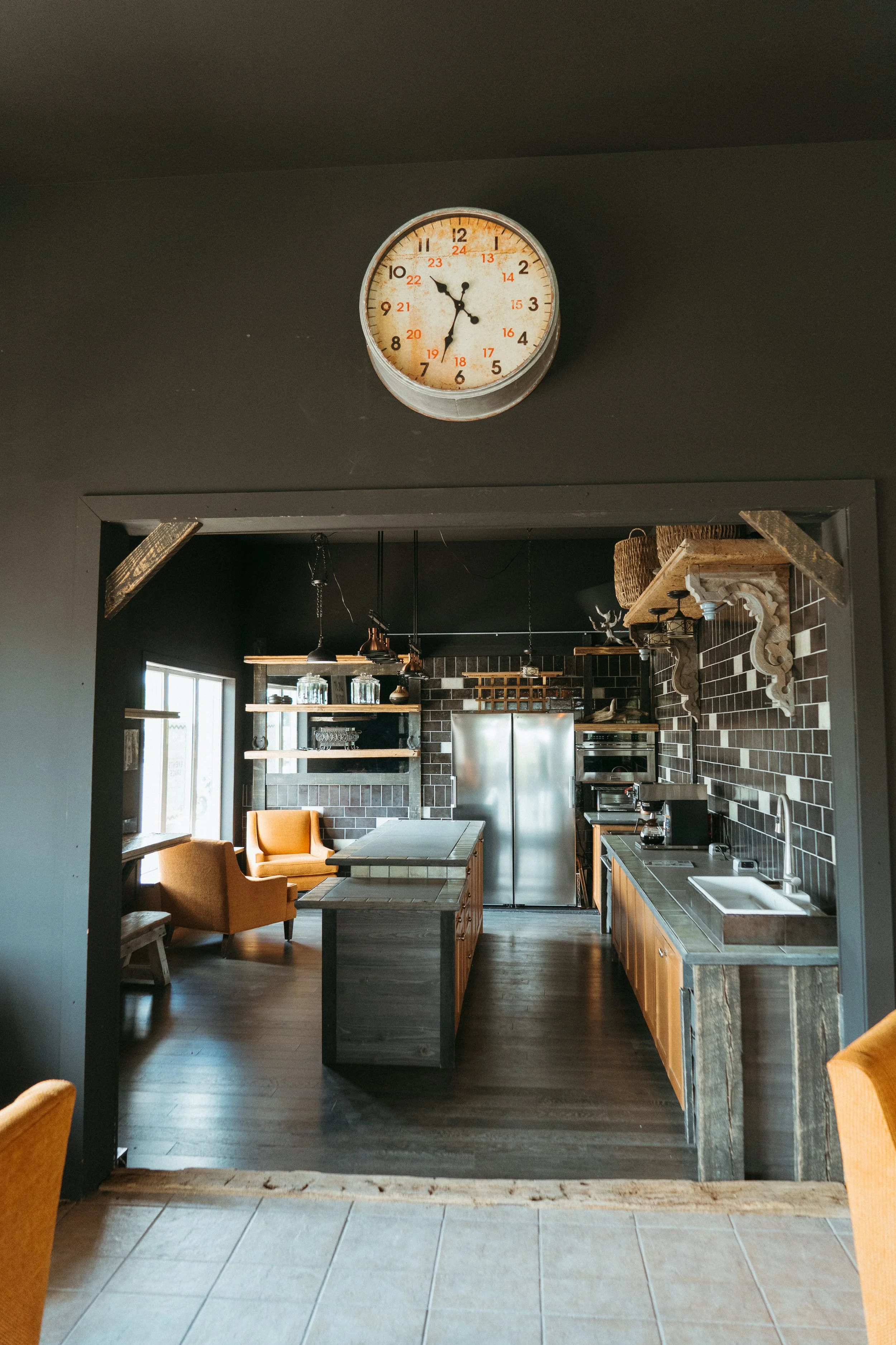 Interior of a modern kitchen with dark walls, wooden cabinetry, stainless steel appliances, and a dining area with orange chairs. A round wall clock shows the time as 5:31.