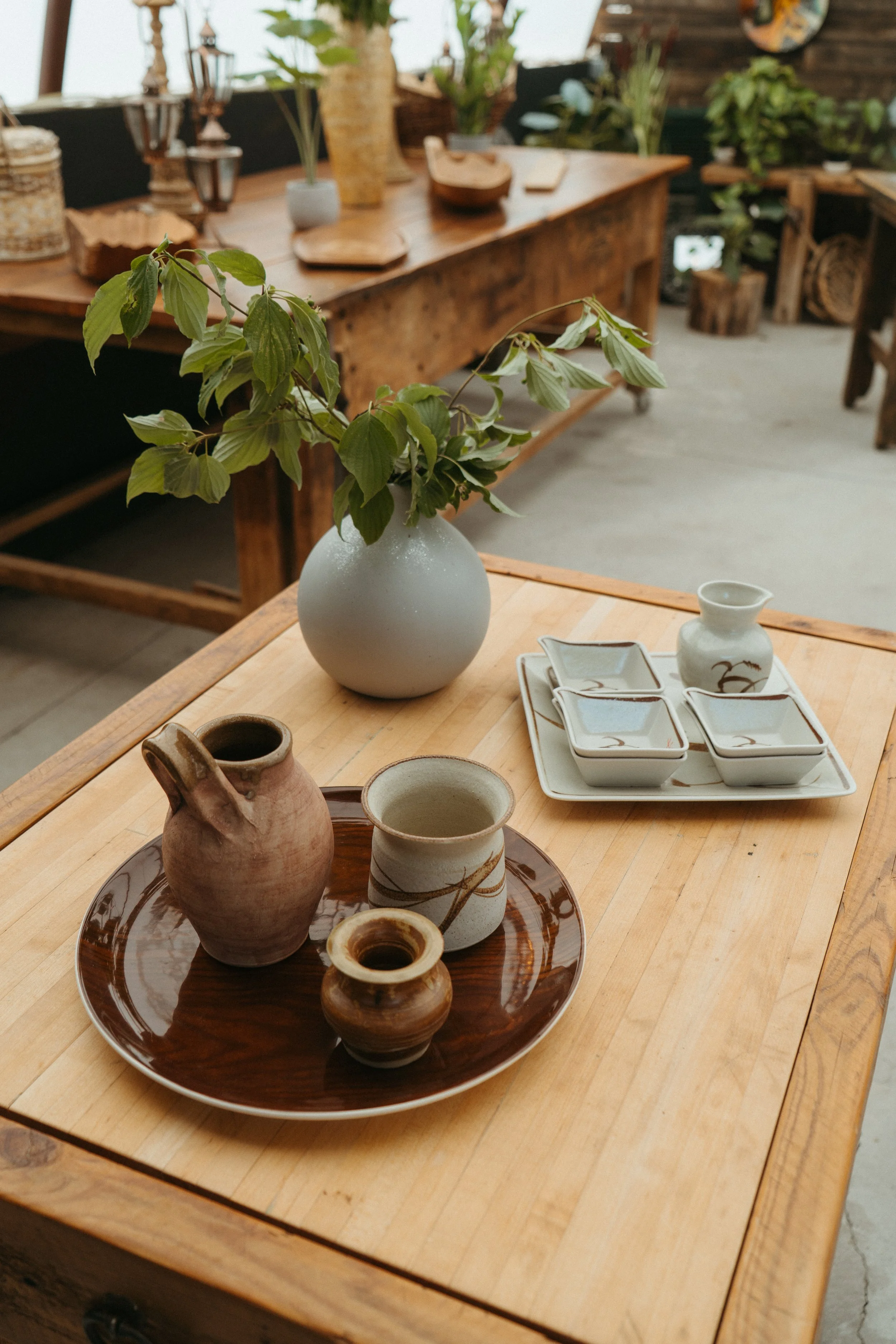 A wooden table with various ceramics and pottery, including vases, cups, and a tray, along with a decorative branch, in a rustic setting with more decor and furniture in the background.