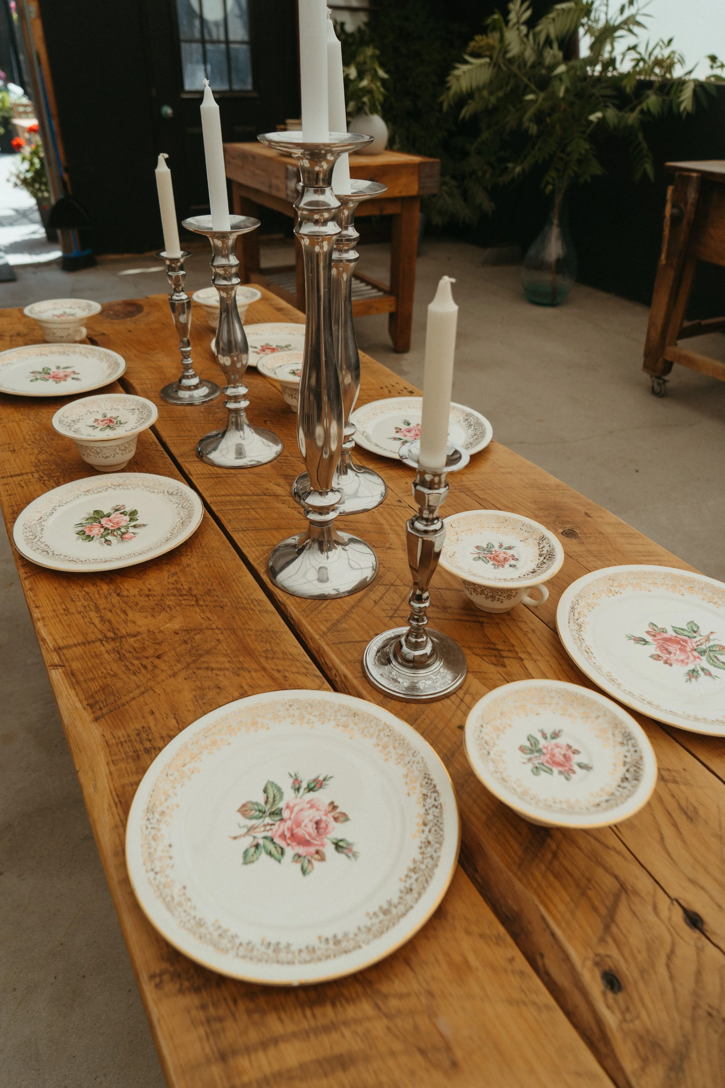 A wooden table set with vintage floral china plates, small bowls, and tall glass candlesticks with white candles, in an indoor or semi-outdoor setting with plants in the background.