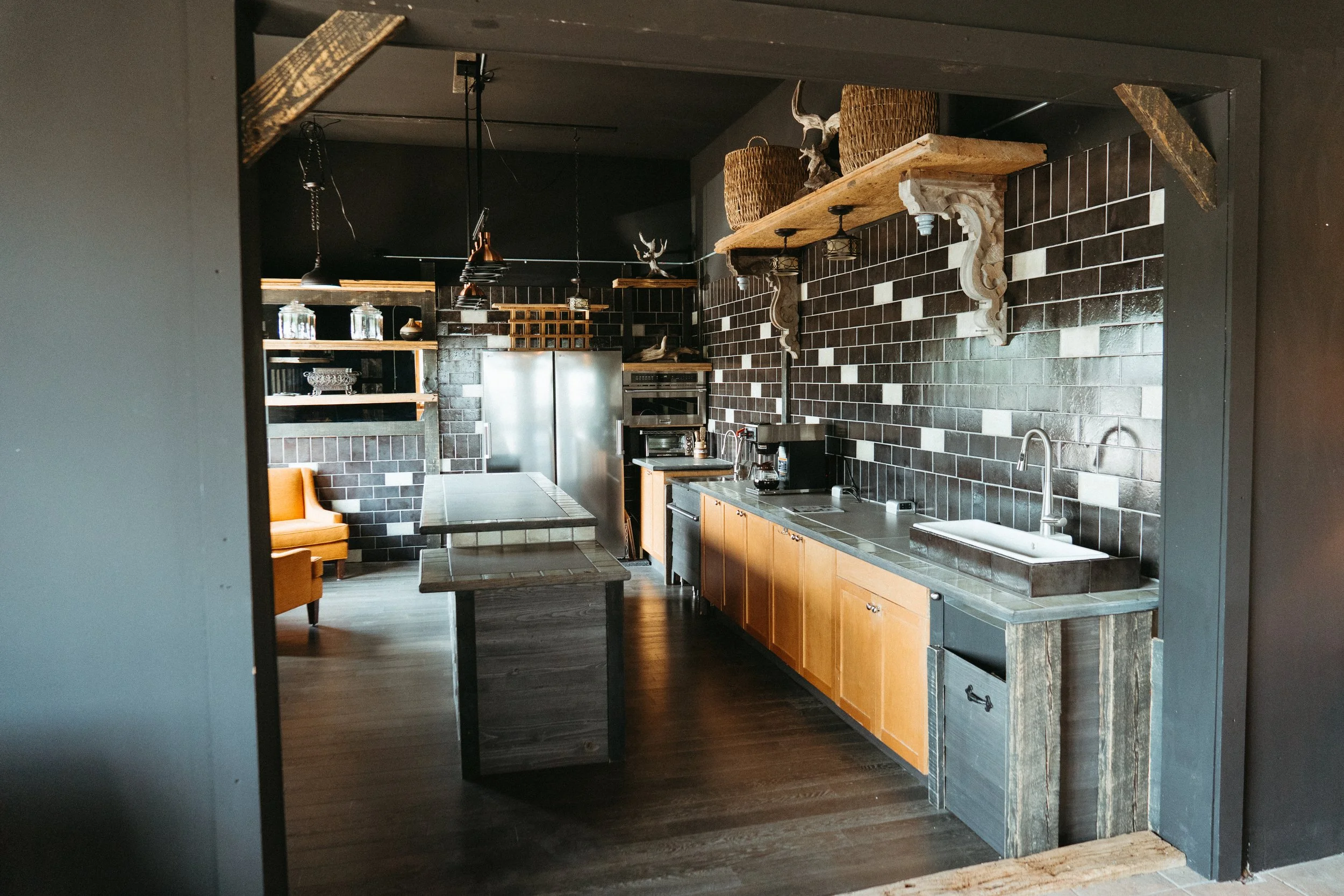 A rustic kitchen with black tiled walls, wooden cabinets, and open wood shelves decorated with antler and natural decor. There is a stainless steel refrigerator, a sink under a window, and hanging black and copper pendant lights.