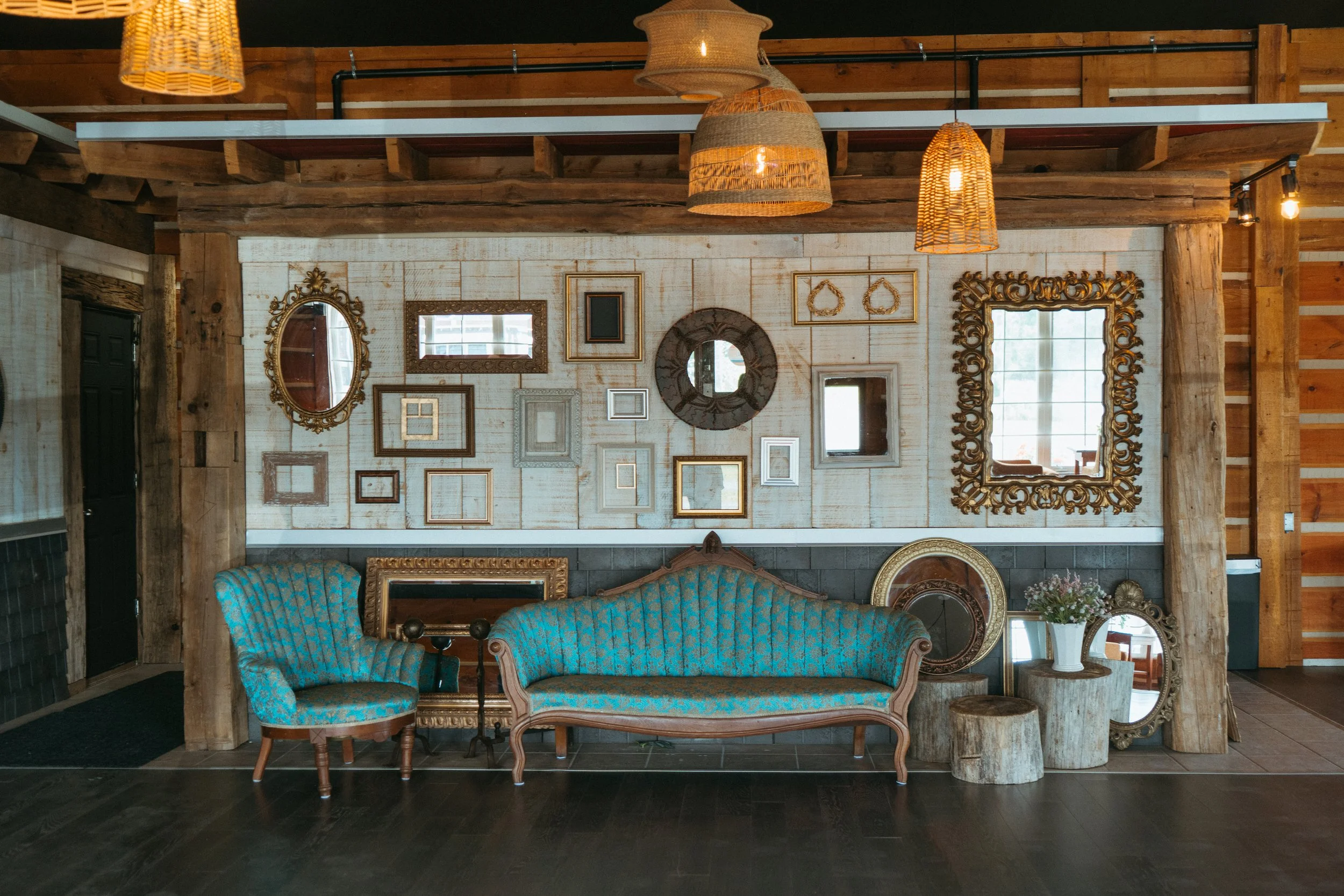 Interior of a rustic room with wooden beams, a wall decorated with various picture frames, a blue upholstered vintage sofa, and a small matching armchair. Hanging wicker lamps illuminate the space, with a window reflected in the wall mirrors.