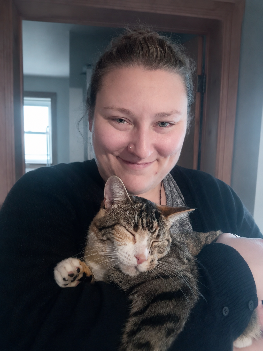 A woman with light skin, light brown hair tied in a bun, and a nose piercing, holding a tabby cat with closed eyes and relaxed expression inside a home.