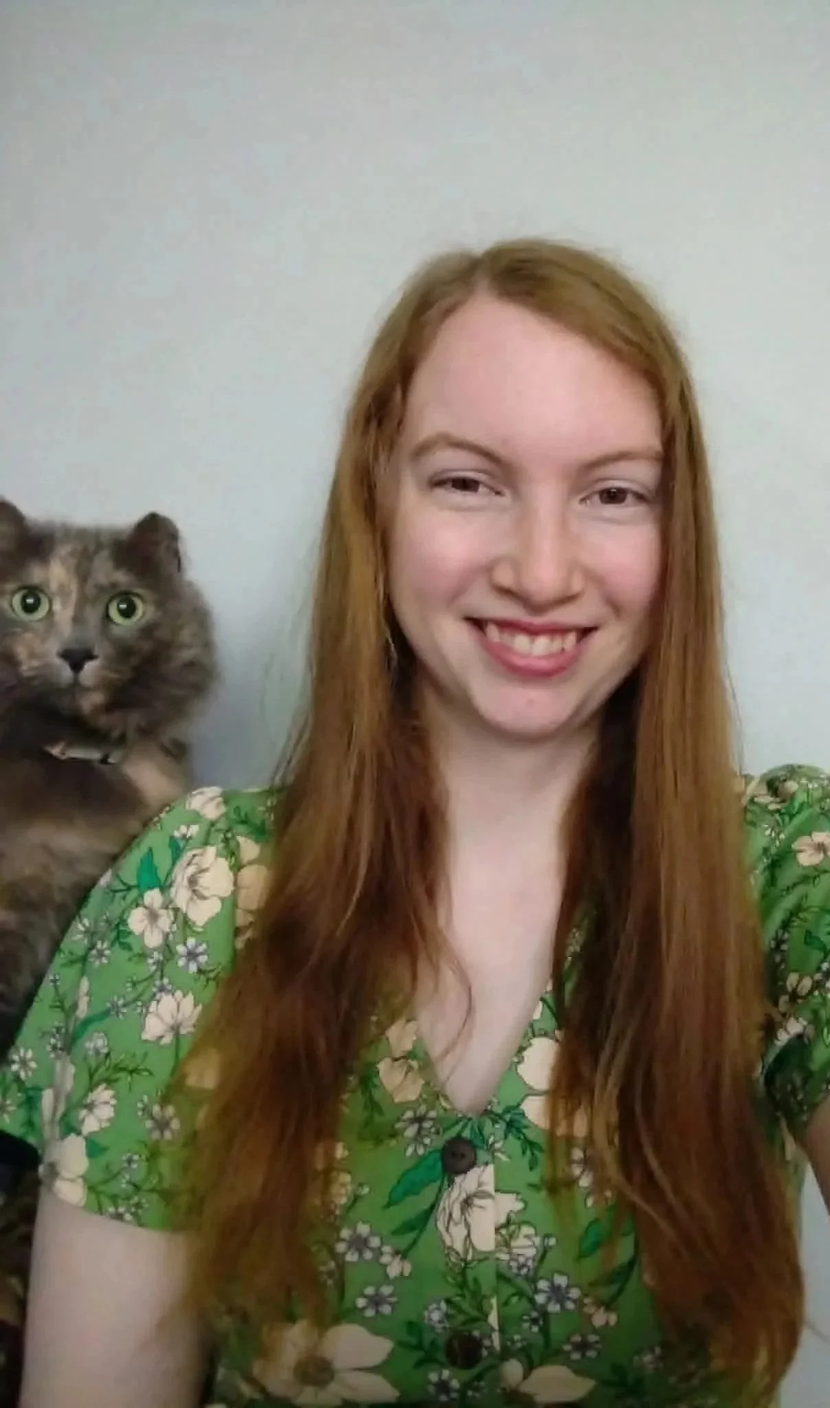 A young woman with long red hair smiling and wearing a green floral blouse, holding a tabby cat with green eyes against a plain white wall background.