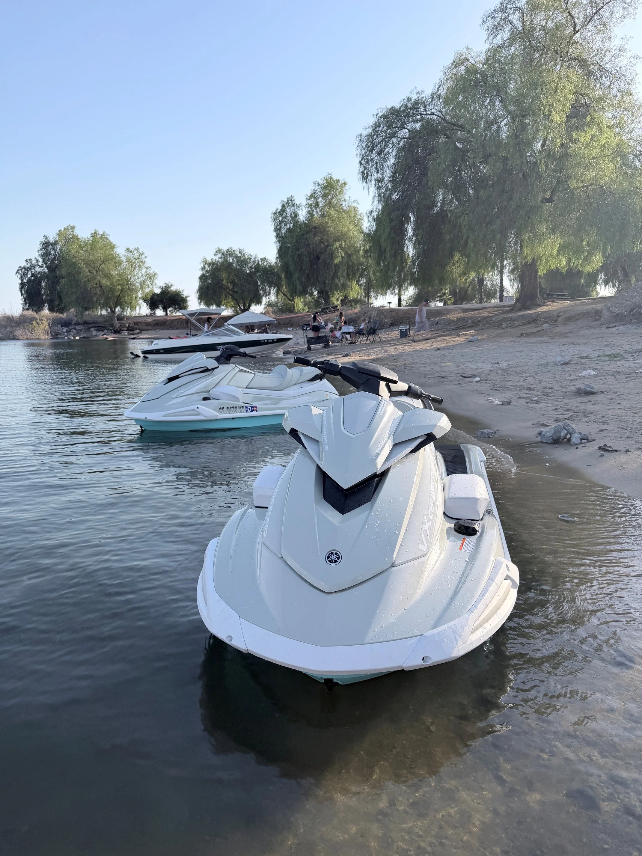 White jet ski in the water near a sandy shore with trees and people having a gathering in the background.