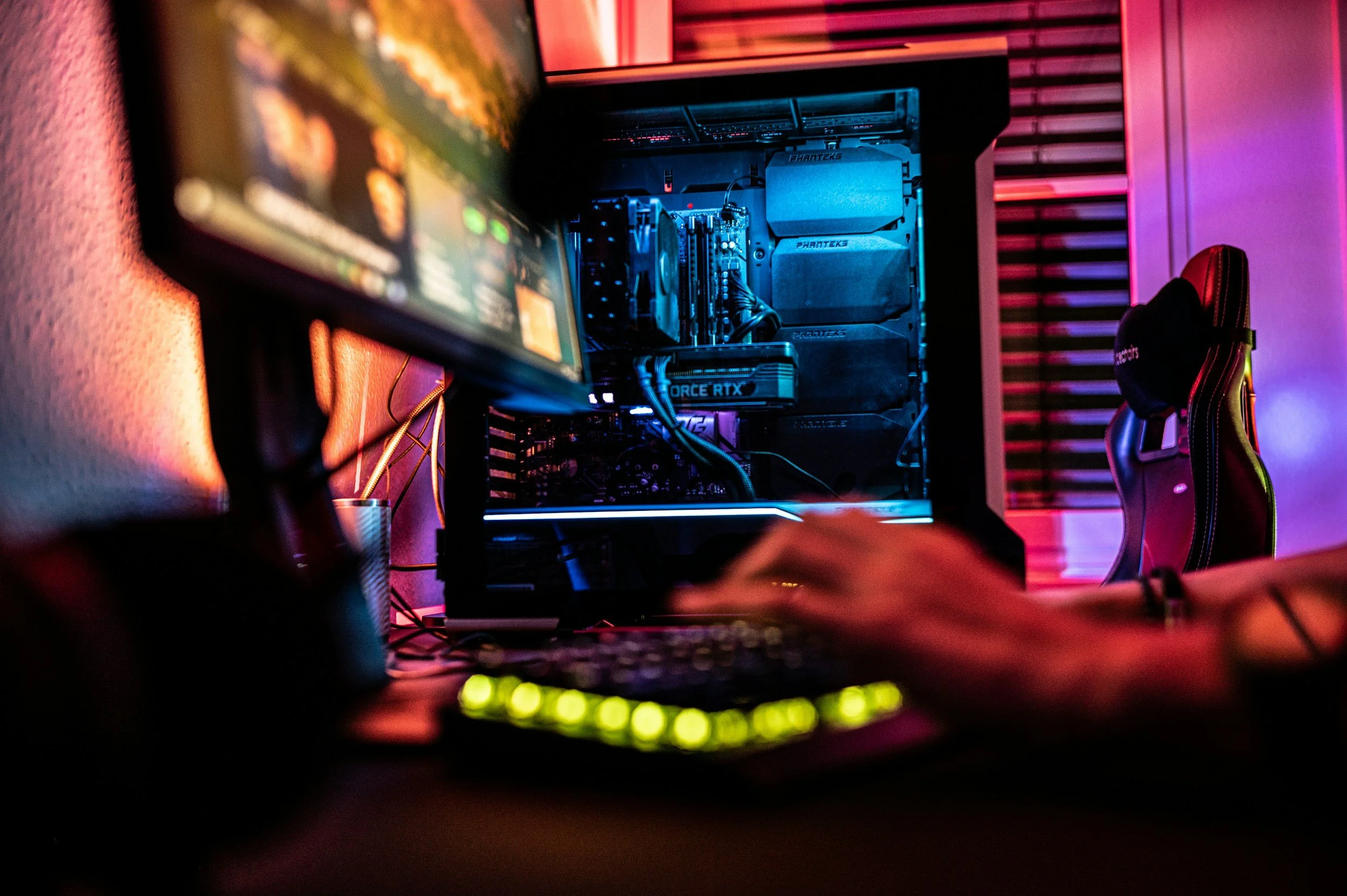 A gaming setup with a monitor, computer tower with blue LED lights, gaming chair, and colorful ambient lighting in a room.