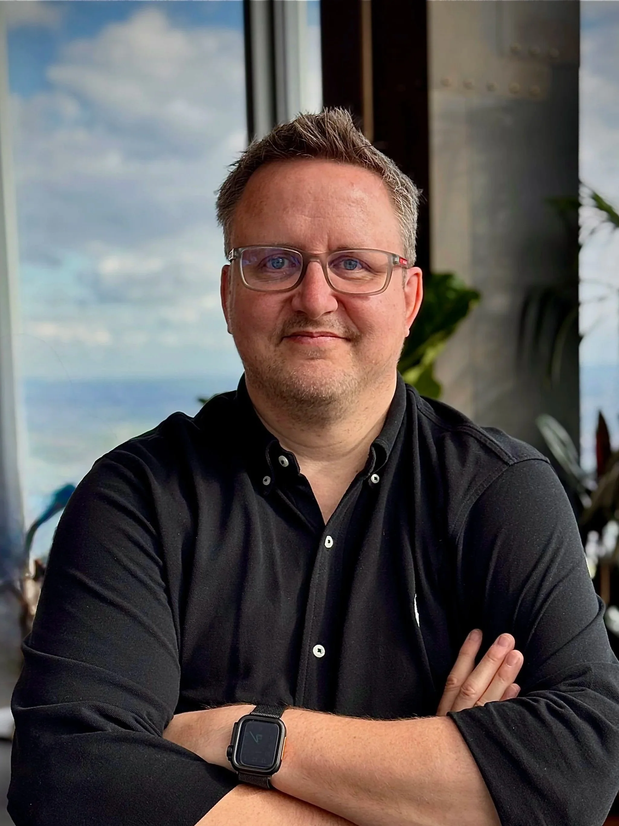 A man with glasses and short, light brown hair sitting at a table with a mountain view outside a large window. He is wearing a black shirt and a smartwatch, with arms crossed on the table.