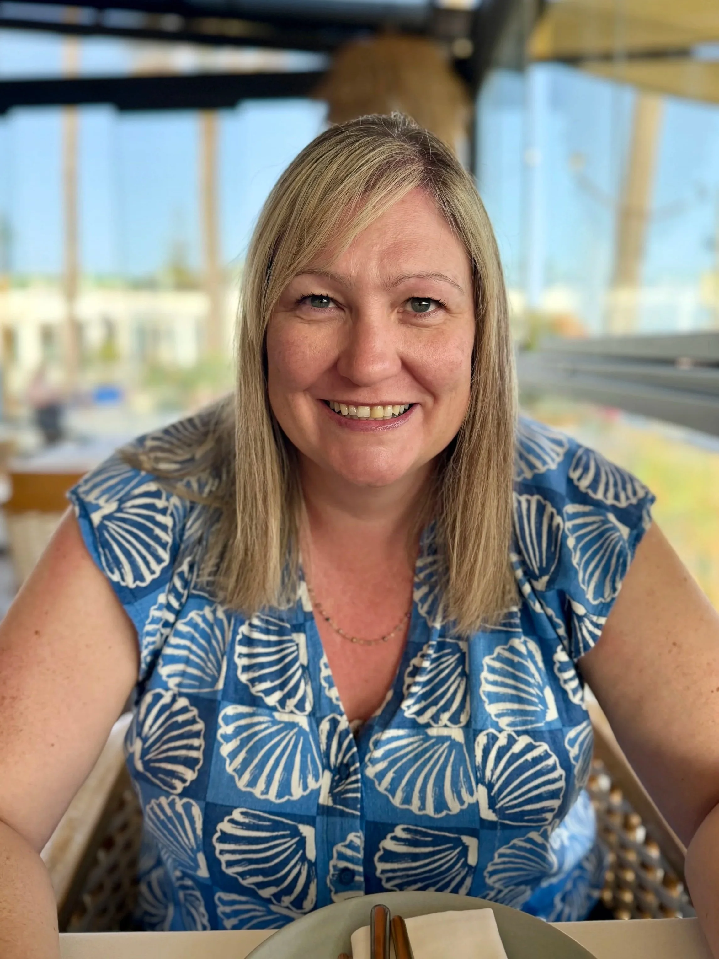 A smiling woman with long blonde hair wearing a blue patterned blouse, sitting at a table with chopsticks and a napkin, in a well-lit restaurant with large windows in the background.