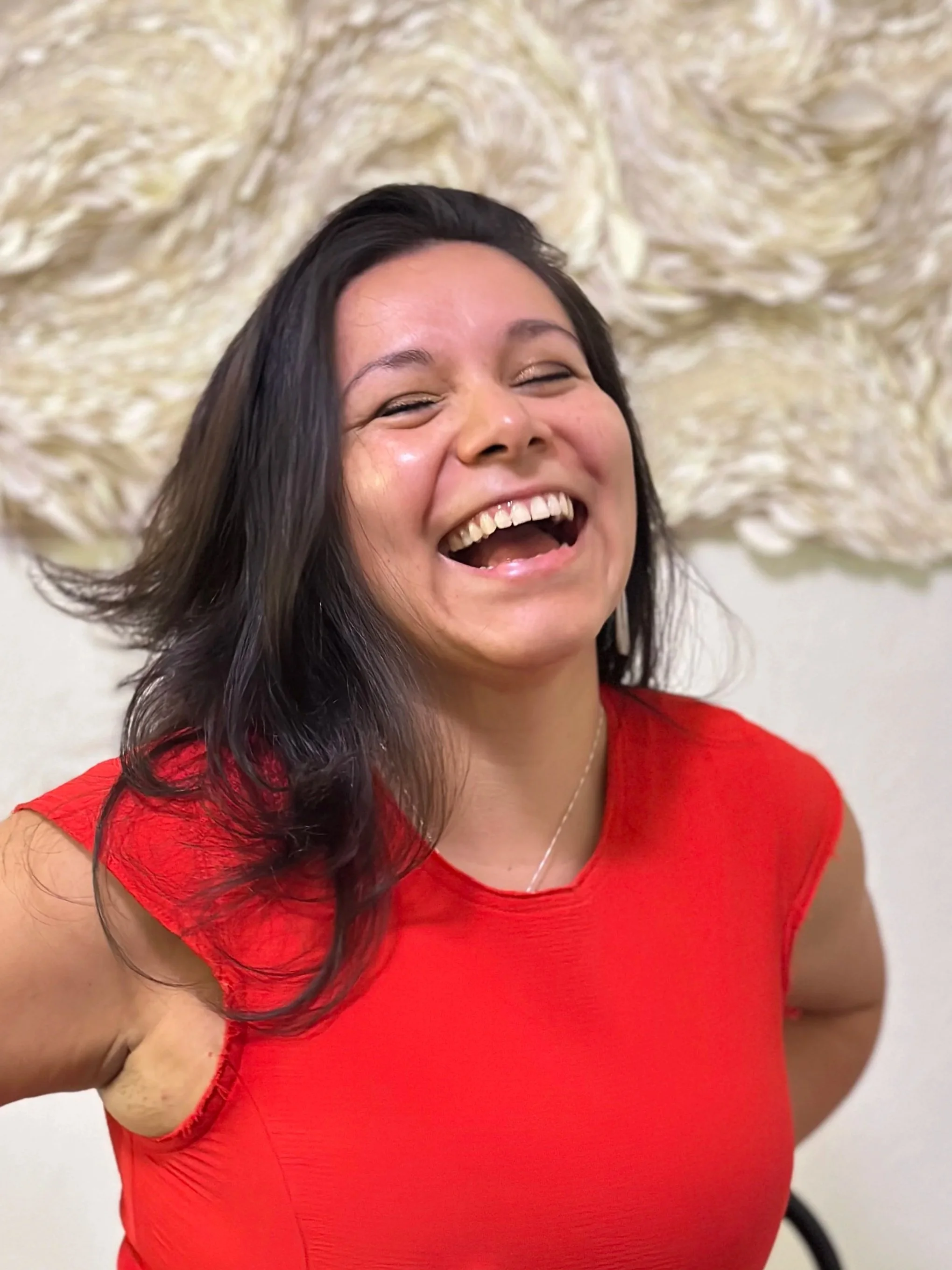 A woman with dark hair, wearing a red top, smiling widely with her eyes closed, against a textured cream-colored background.