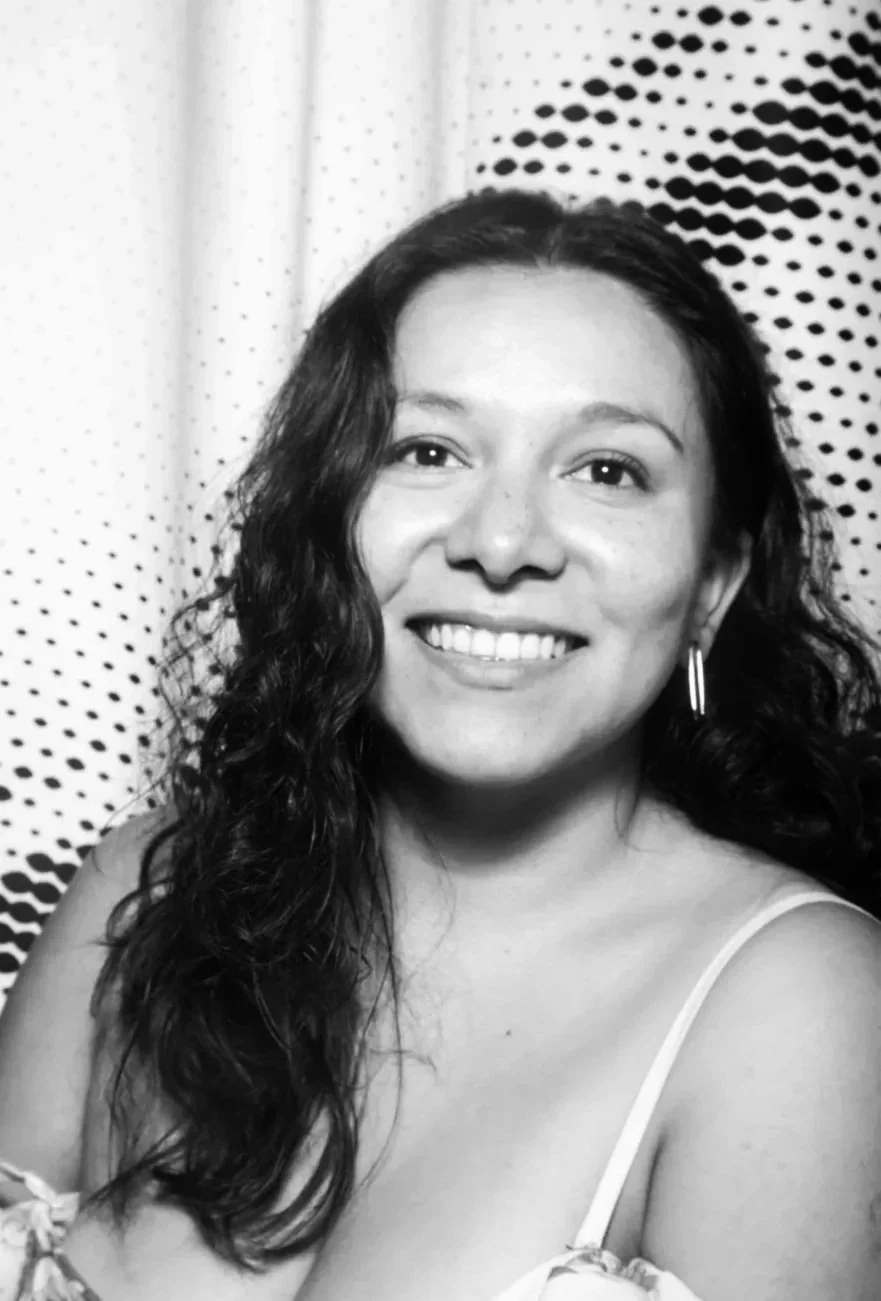 A smiling woman with wavy dark hair, wearing earrings and a thin-strap top, in front of a patterned curtain.