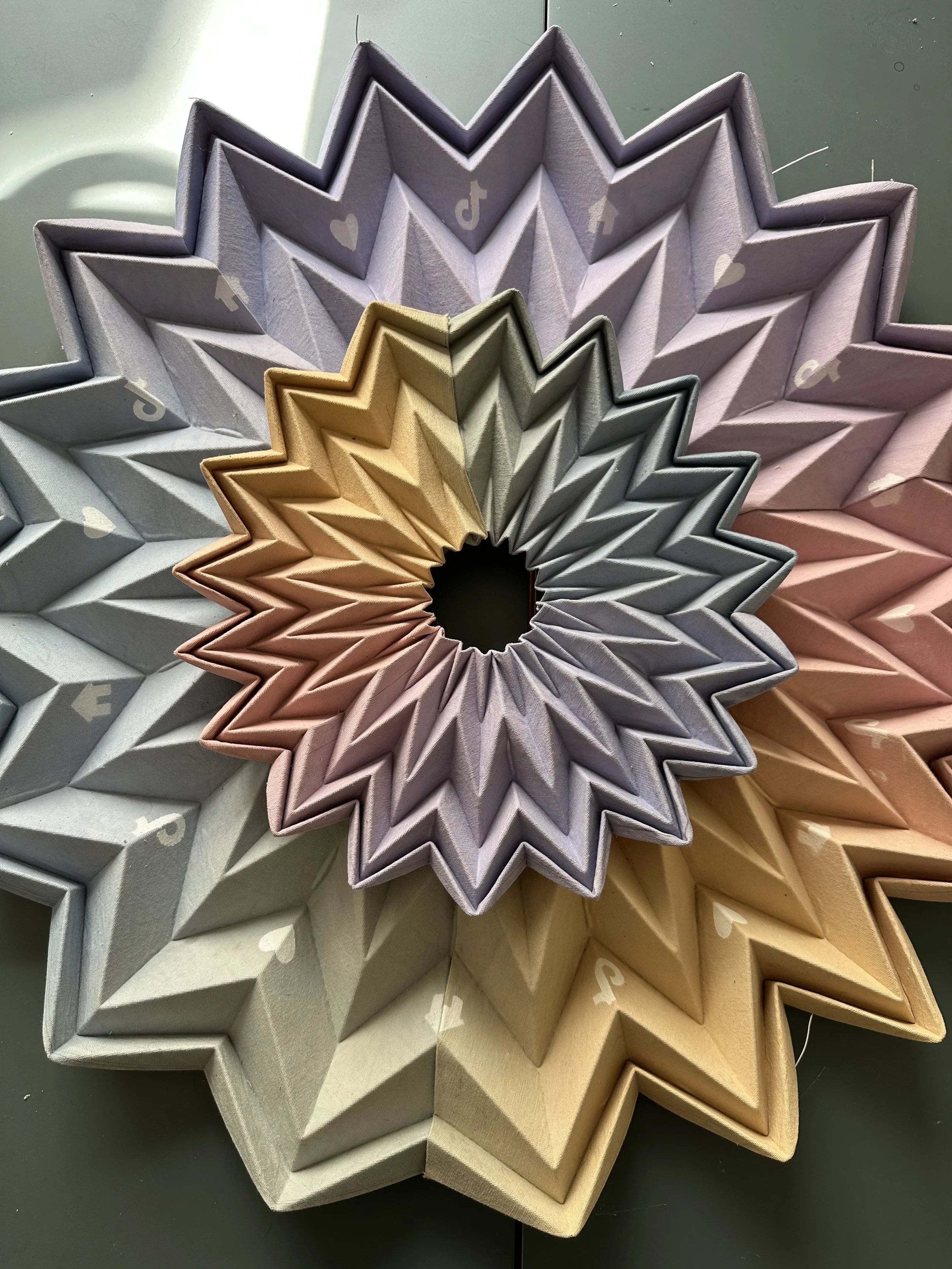Colorful star-shaped paper decorations with heart and arrow symbols, overlapping each other in pastel shades of purple, gray, pink, peach, and yellow, arranged in a circular pattern.