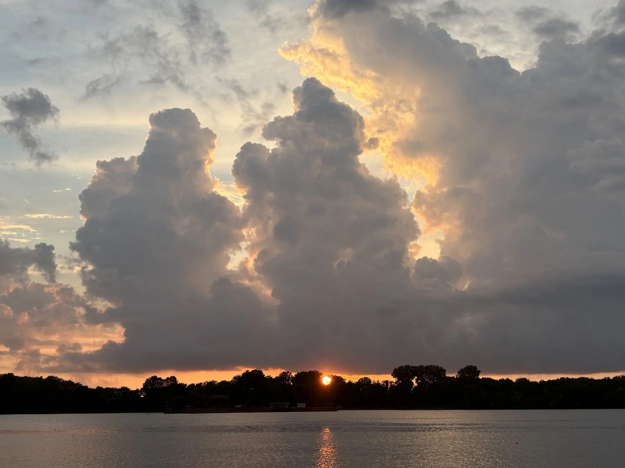 Sunset over a lake with dark clouds in the sky, orange and yellow hues glow from the setting sun near the horizon, silhouetted trees along the shoreline.