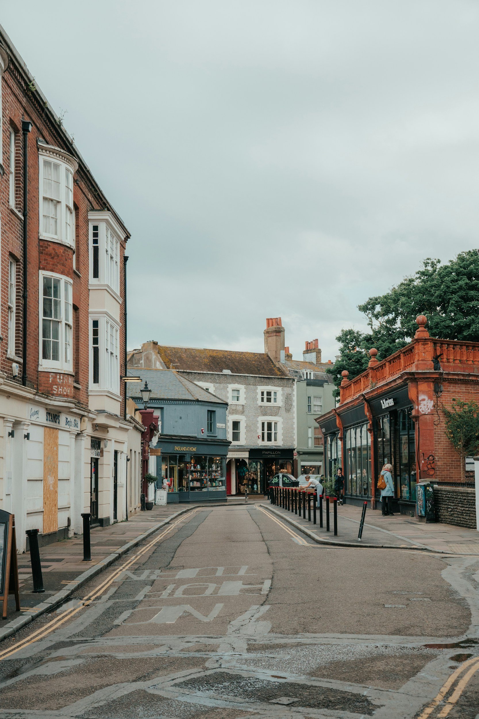 A street scene with shops and pedestrians in Brighton, England. Buildings include brick and painted facades with shop signs, and there are trees and a cloudy sky overhead.
