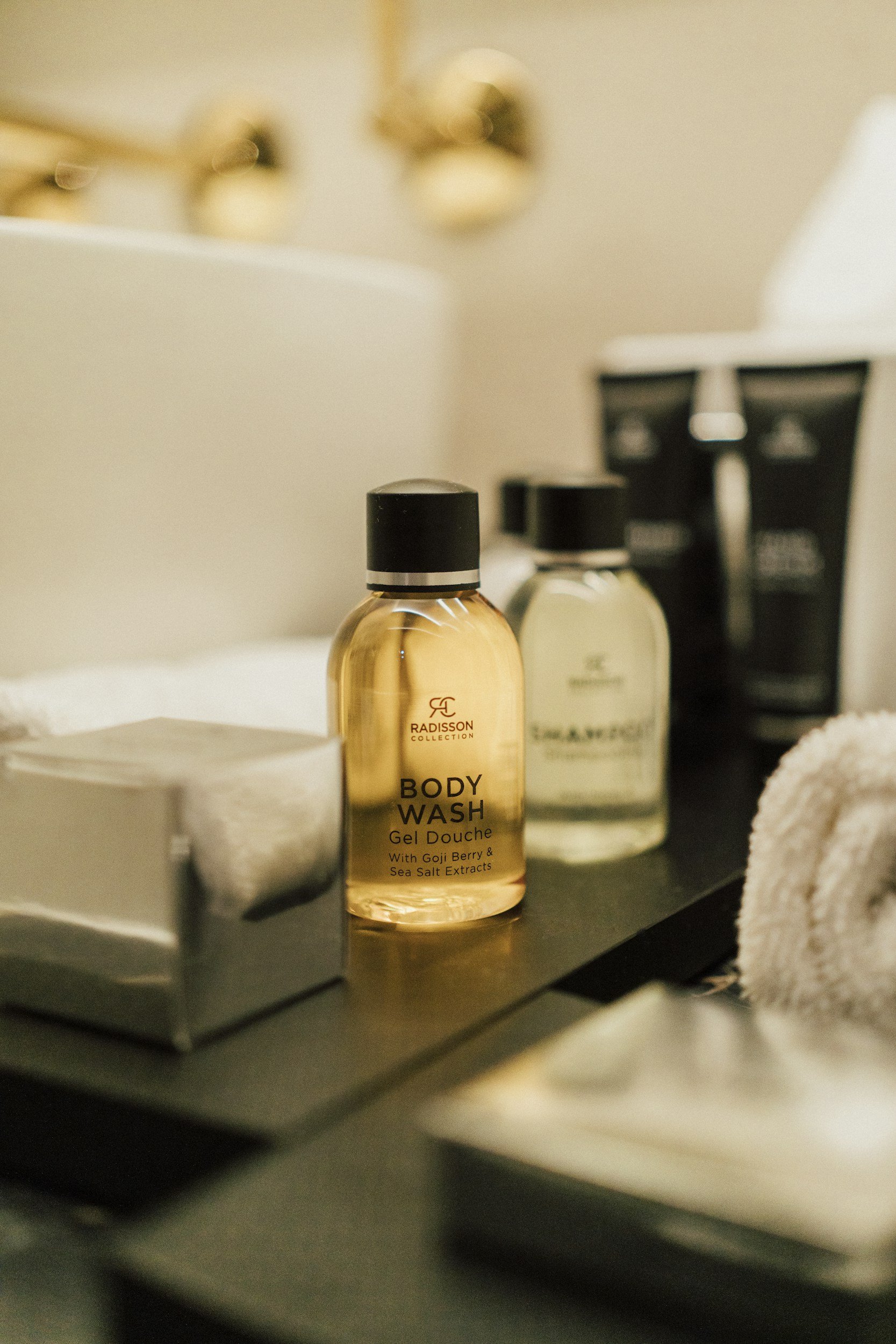 A bottle of Radisson Collection body wash on a dark surface, with a candle, other bottles, and a rolled towel nearby in a bathroom setting.