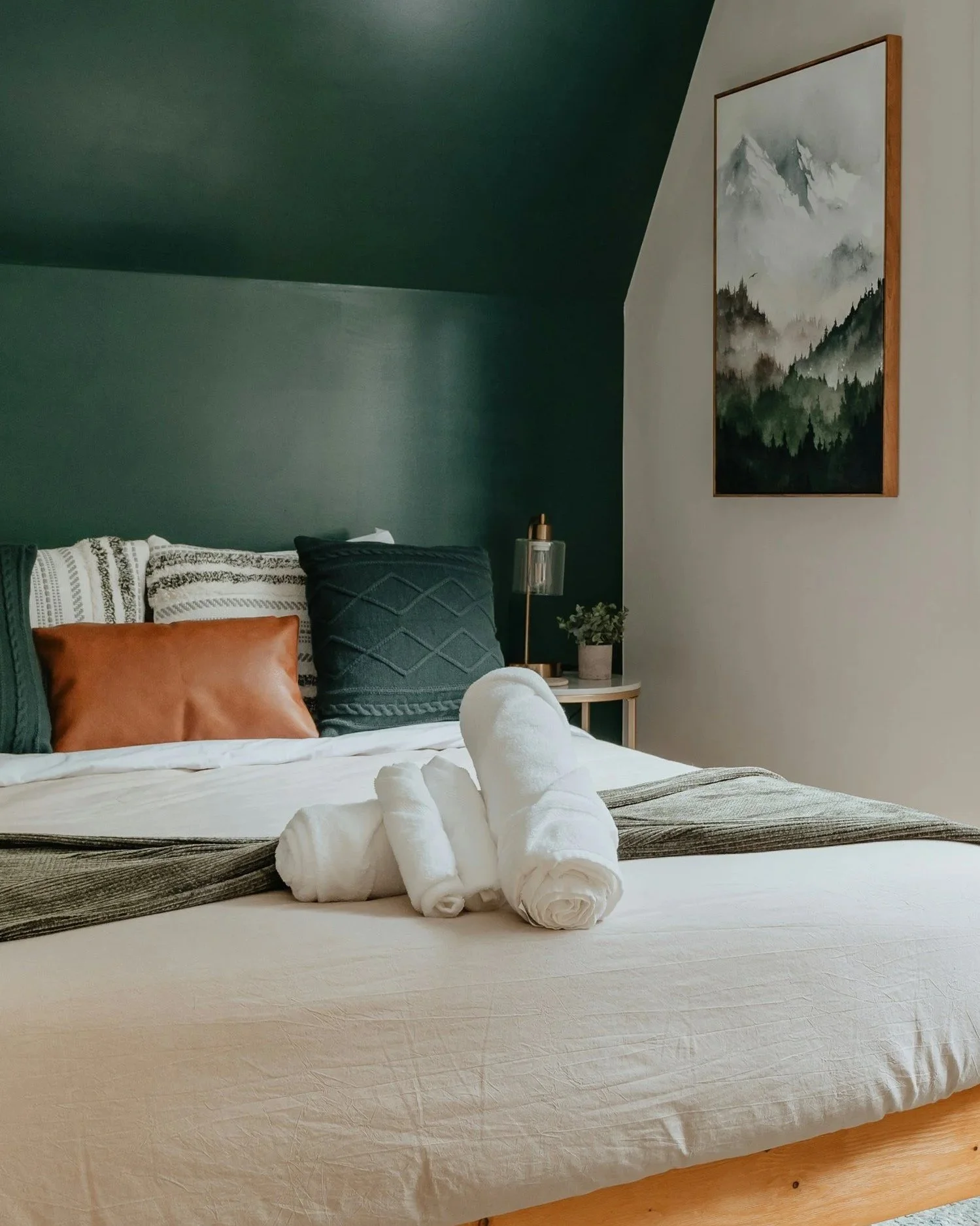 A neatly made bed with rolled white towels on top, decorated with multiple pillows in shades of beige, black, and patterned fabric, and a striped blanket at the foot of the bed. A bedside table with a small plant and a modern lamp is visible. A mountain landscape painting hangs on the wall in the background.