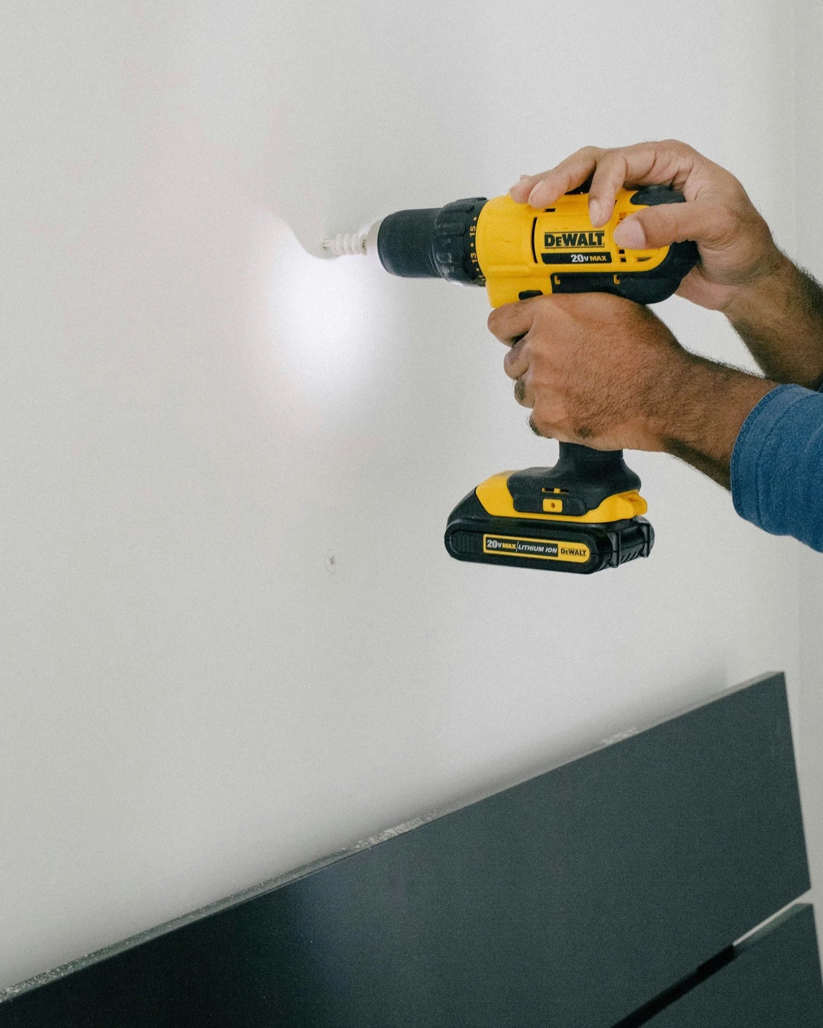 Hand holding a yellow DeWALT cordless drill, drilling into a white wall, with a black battery pack attached at the bottom.