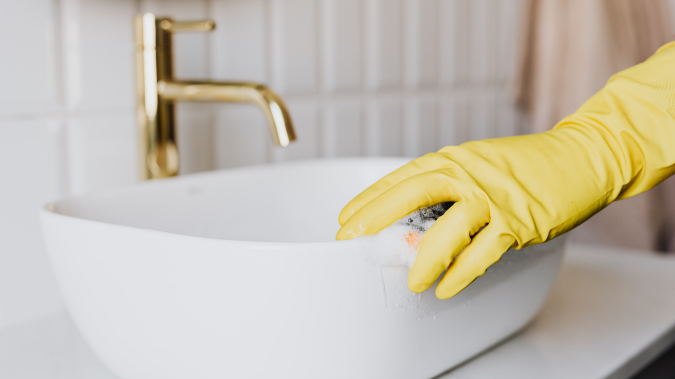 Person wearing yellow rubber gloves washing a white bowl with soap in a kitchen sink.