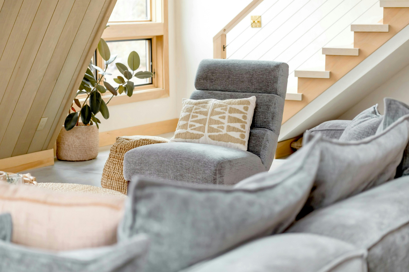 Cozy living room corner with a gray armchair, decorative pillows, a potted plant, and large windows in a modern, wood-accented space.