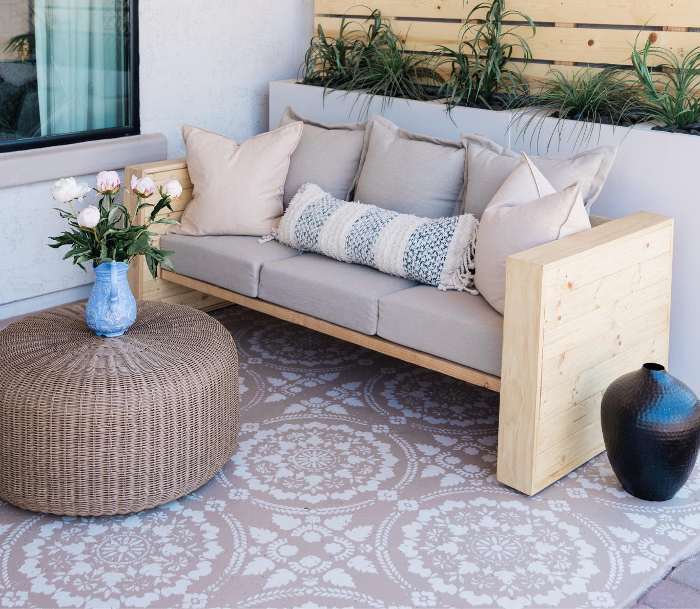 Outdoor patio area with a light-colored wooden framed sofa with beige cushions and a decorative throw pillow, a large round wicker ottoman with a blue vase holding flowers on top, a patterned outdoor rug, a tall black decorative vase, and potted plants on a planter box against a wooden slat wall.