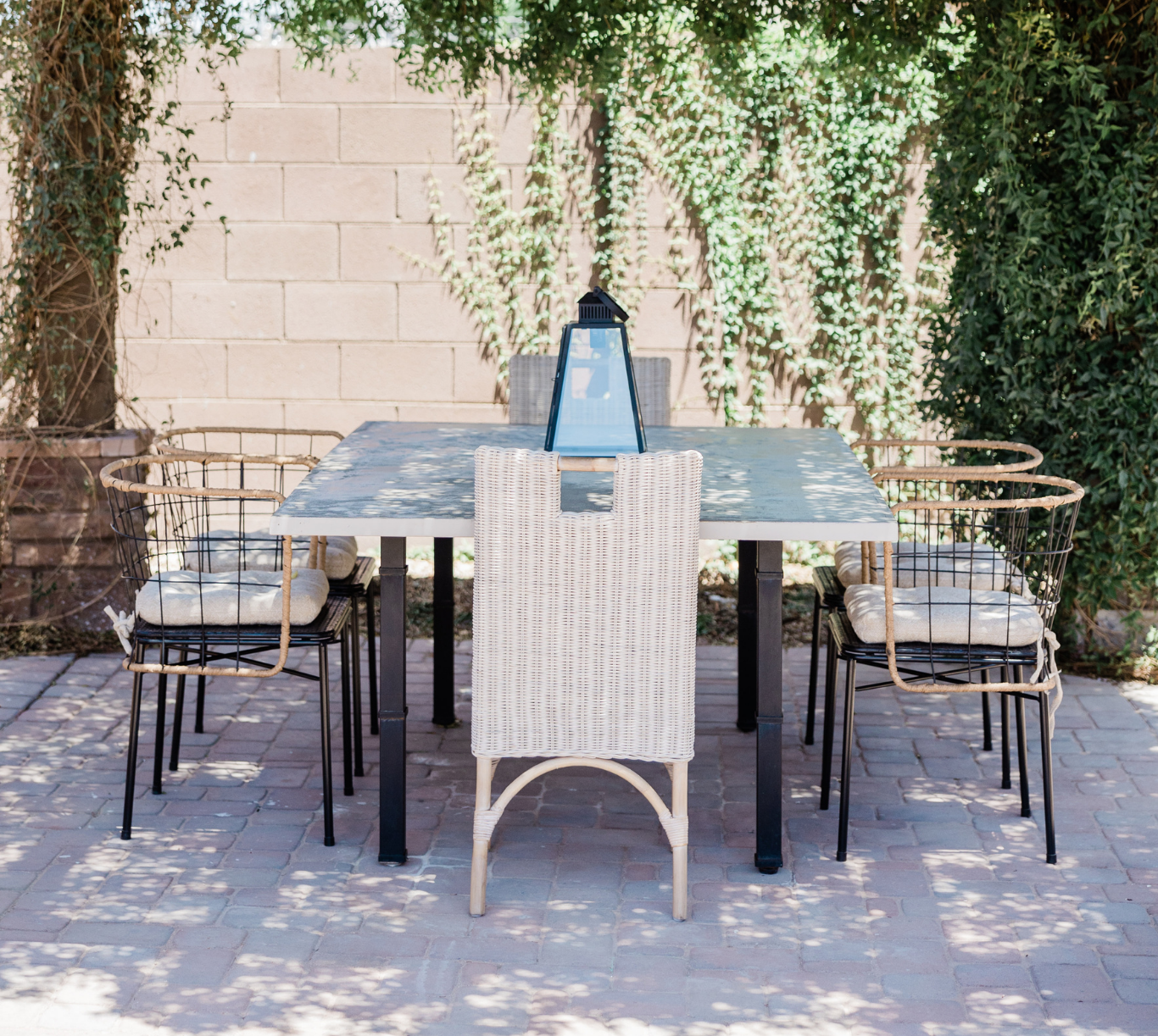 Outdoor patio dining table with six chairs and a blue lantern in the center, surrounded by greenery and brick paving.